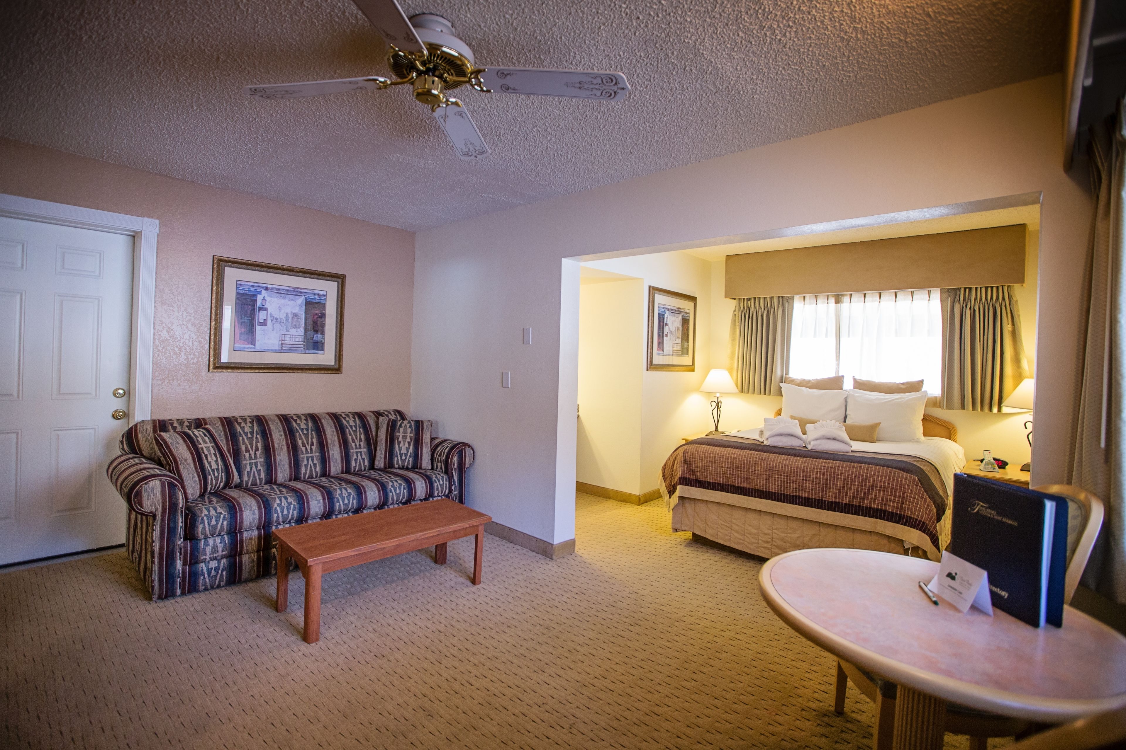 A hotel room with a patterned sofa, a wooden bench, a bed with a headboard, a bedside table with a lamp, and a ceiling fan.