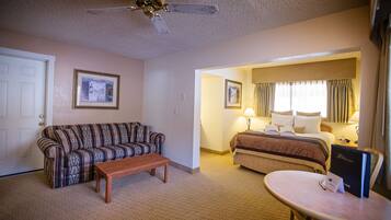 A hotel room with a patterned sofa, a wooden bench, a bed with a headboard, a bedside table with a lamp, and a ceiling fan.