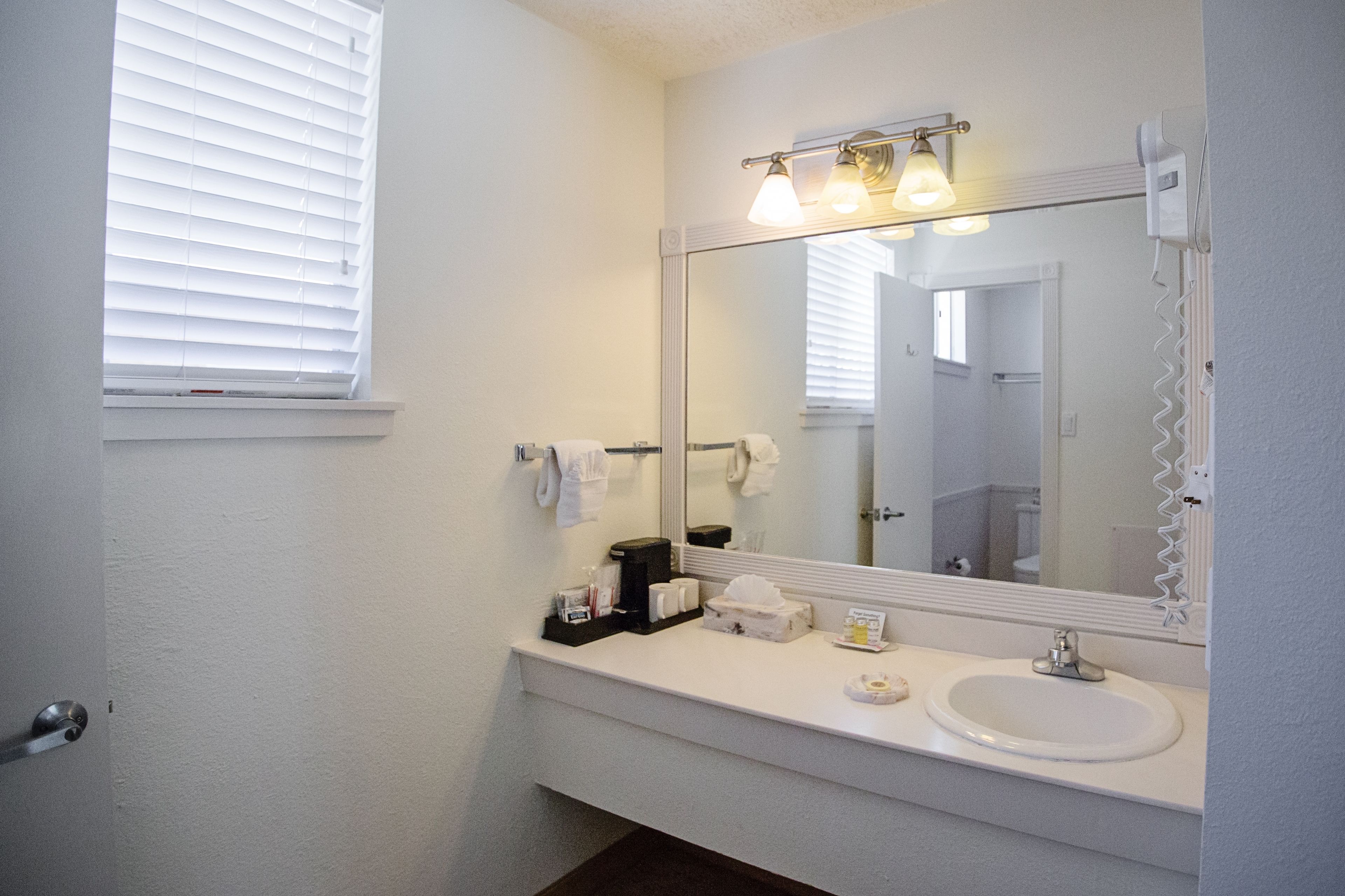 Un baño con lavamanos blanco, un espejo grande y una ventana con persianas.