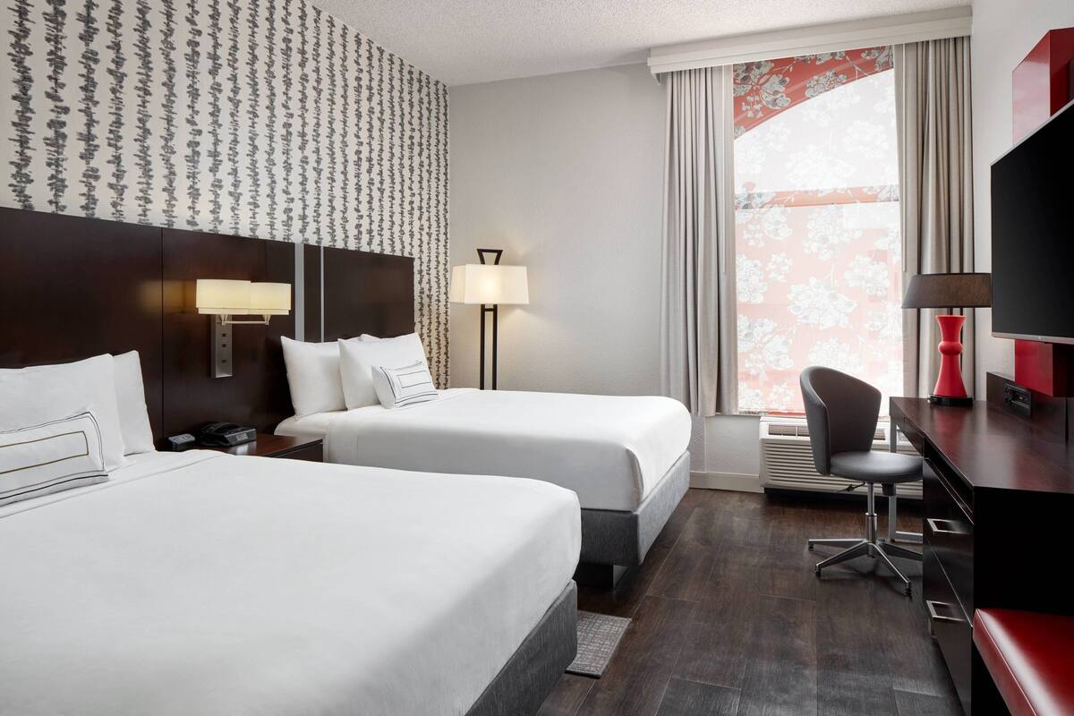 Quarto de hotel com duas camas, uma escrivaninha, uma cadeira, uma televisão e uma janela com cortinas.