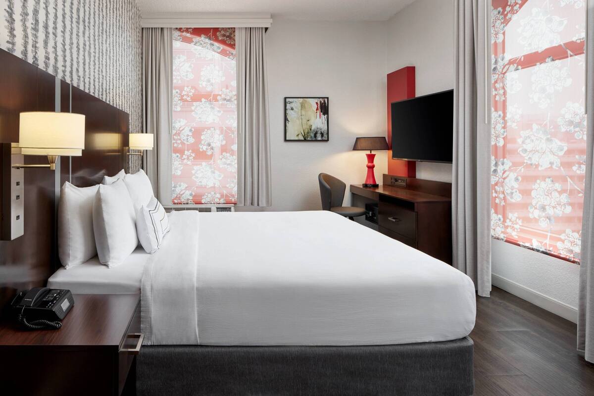 Quarto de hotel com uma cama grande, luminárias de cabeceira, uma escrivaninha com telefone, televisão e luminária de mesa.