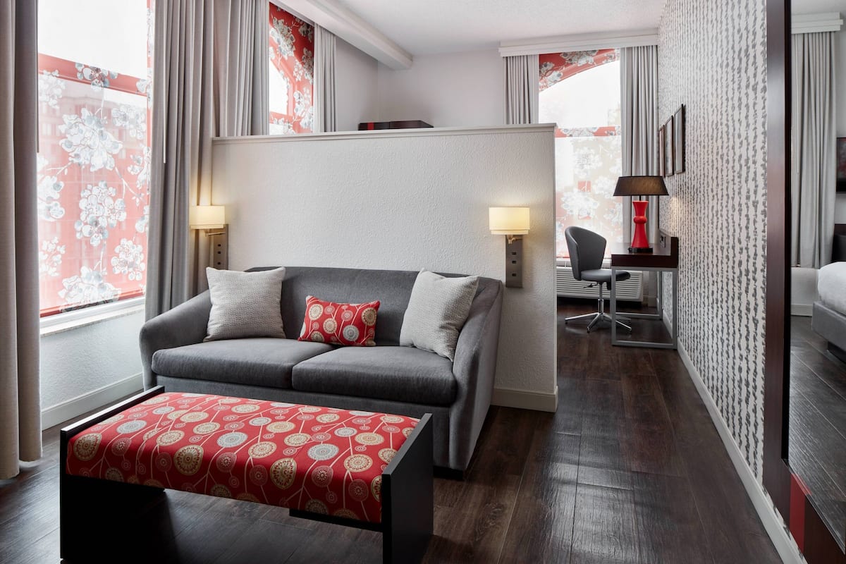 Quarto de hotel moderno com um sofá cinzento, um banquinho estampado em vermelho, uma escrivaninha com luminária e uma cadeira.
