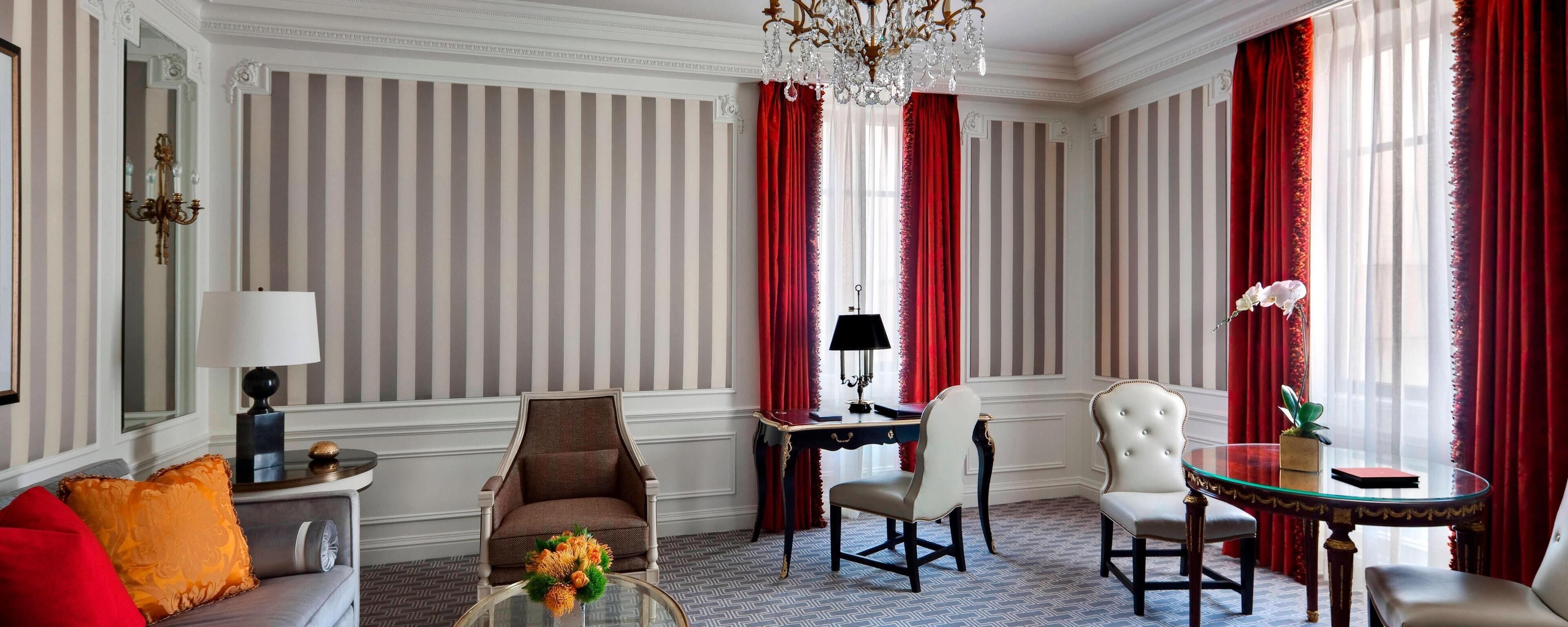 A room with striped wallpaper, red curtains, a chandelier, a sofa, a table, and a chair.