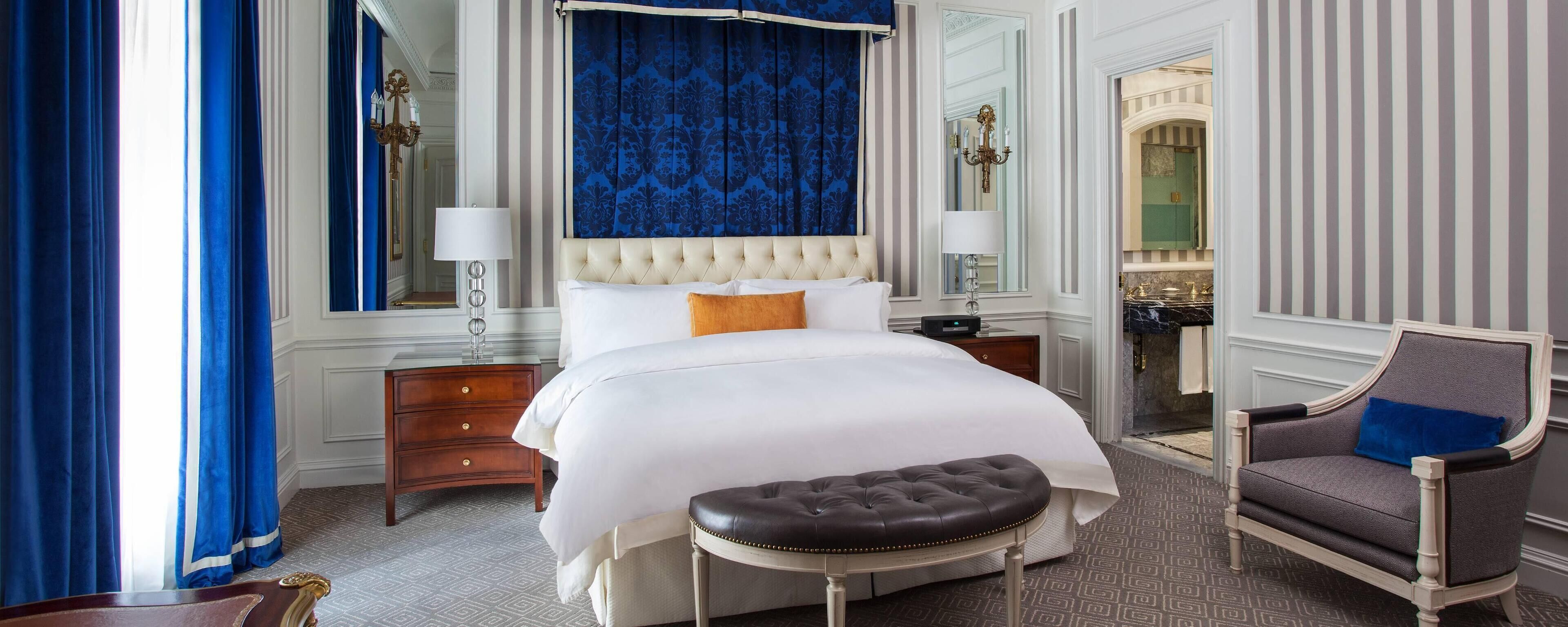 A hotel room with a large bed, a chair, a nightstand, and a bathroom visible through an open door.