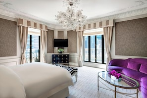 A hotel room with a large bed, a purple sofa, a glass coffee table, a TV, and a view of the city.