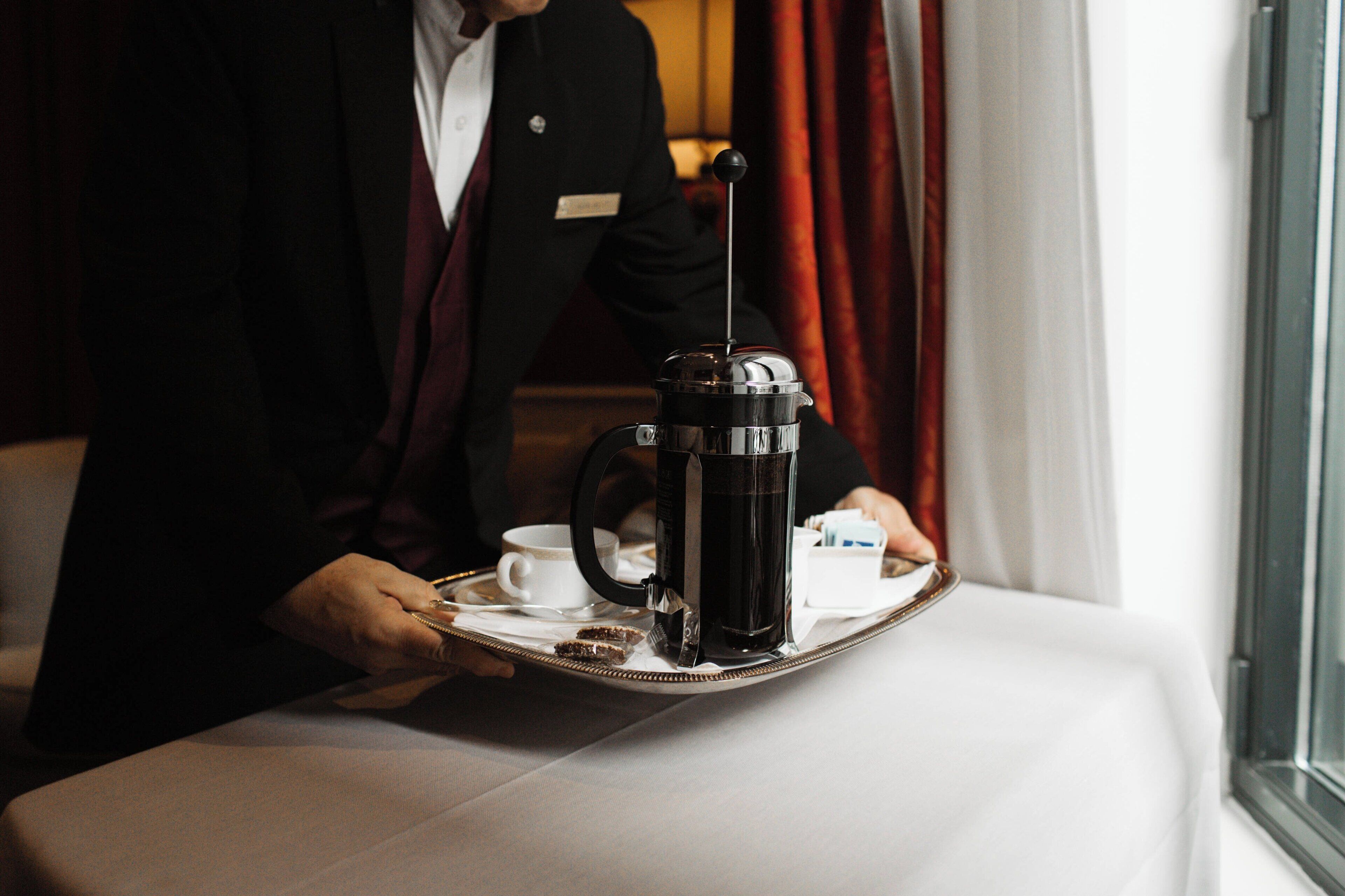 A person in a black suit holding a tray with a coffee maker, a cup, and a box of tissues.