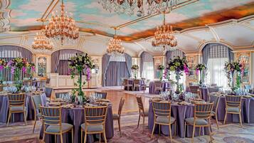 A grand ballroom with ornate chandeliers, elegant round tables, and floral centerpieces.