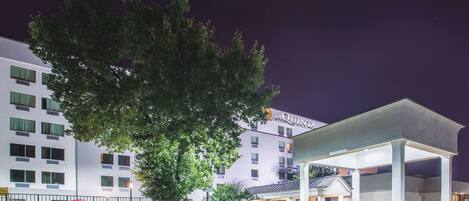 Hotel com área externa coberta, uma grande árvore e fachada bem iluminada.