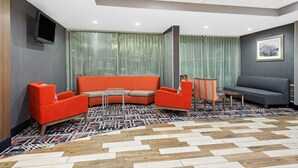 Un área de descanso moderna con alfombra estampada, sofás naranja y grises, una mesita y un televisor montado en la pared.