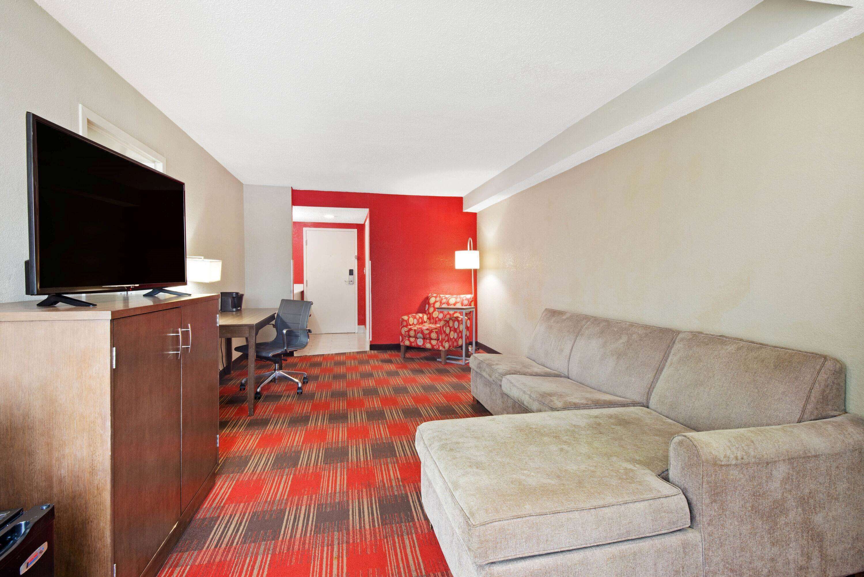 A hotel room with a flat-screen TV, a wooden cabinet, a grey sofa, a red carpet, a desk with a chair, and a red accent wall.