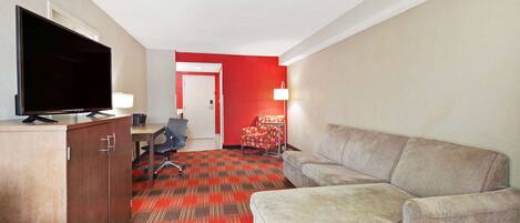A hotel room with a flat-screen TV, a wooden cabinet, a grey sofa, a red carpet, a desk with a chair, and a red accent wall.