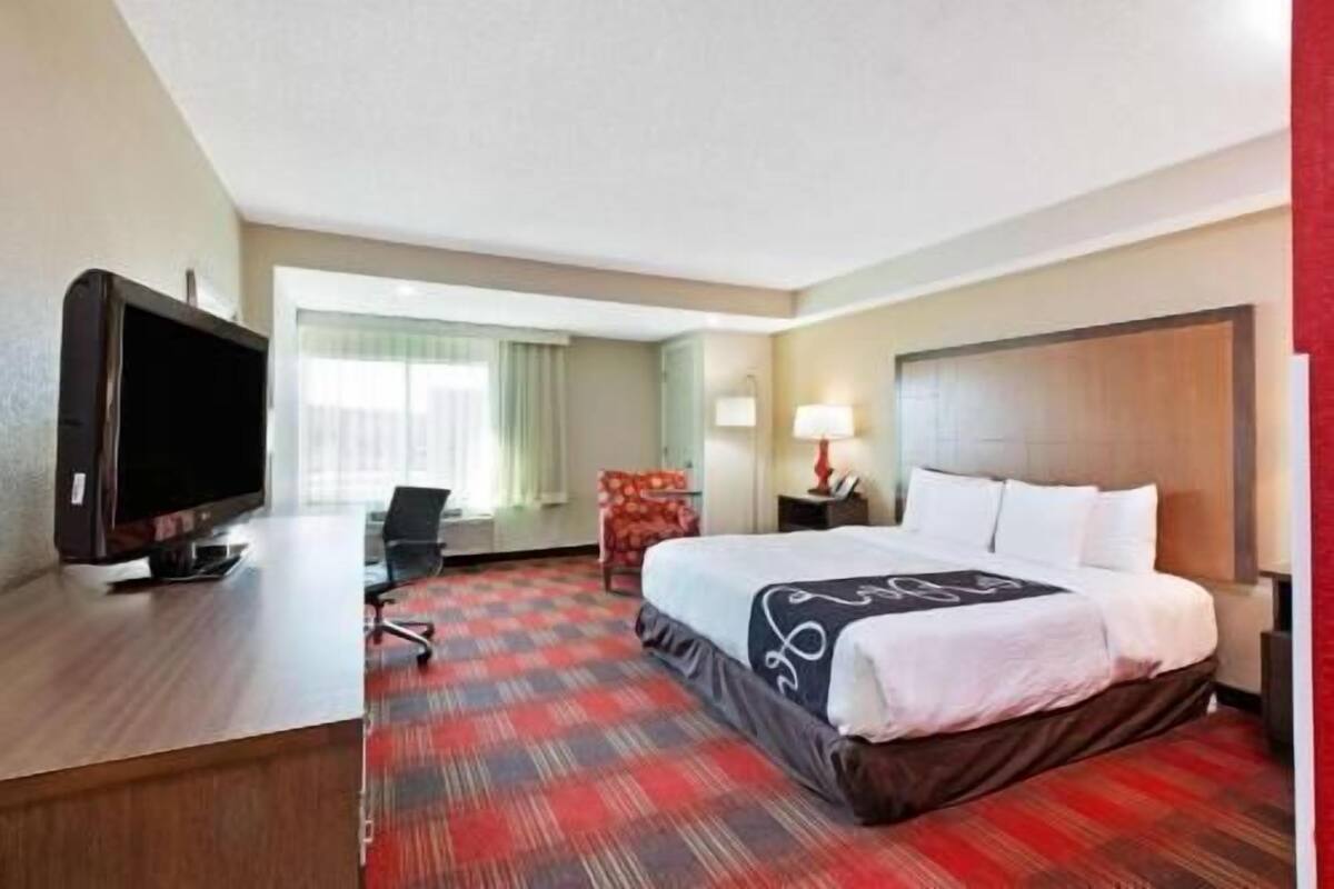 A hotel room with a bed, a desk with a TV, a chair, a lamp, and a window with curtains.