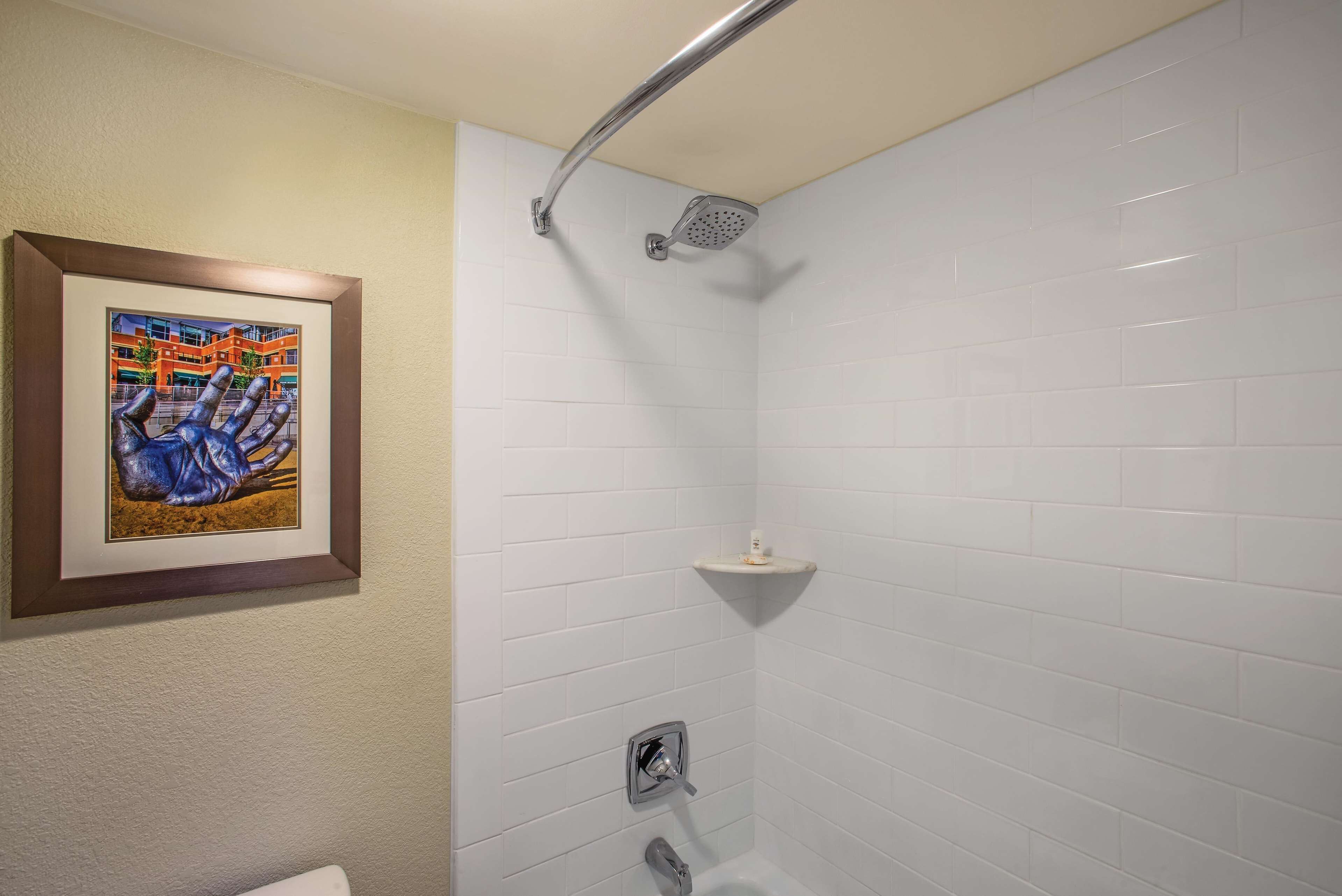 Une salle de bain avec des murs recouverts de carreaux blancs, une œuvre d’art encadrée, une pomme de douche et une petite étagère.