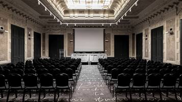 Een congreszaal met rijen stoelen gericht op een groot scherm, een versierd plafond en een kamerbreed tapijt met patroon.