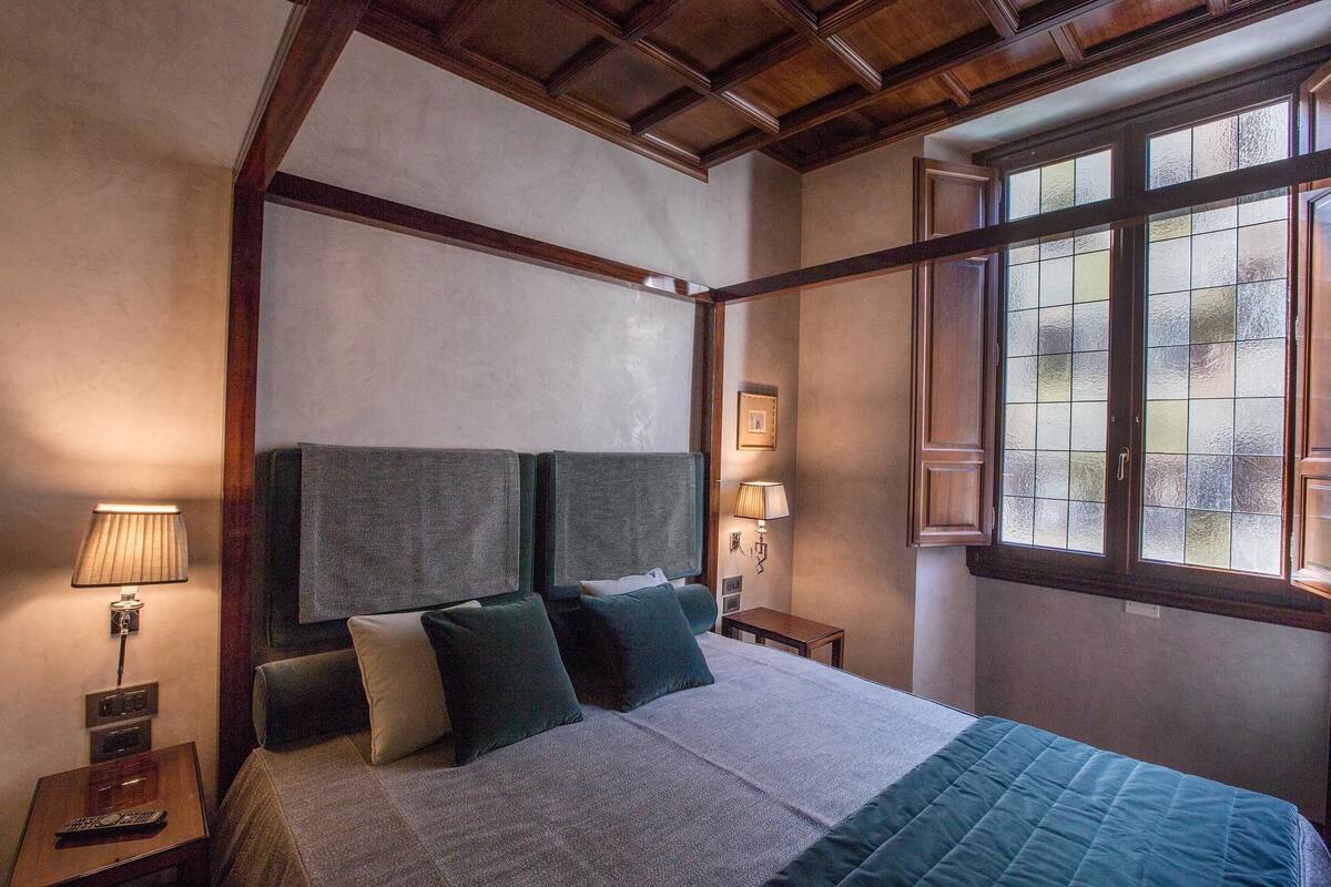 Ein Hotelzimmer mit einem Himmelbett, einem Nachttisch mit Lampe, einem Fenster mit Milchglas und einer Holzbalkendecke.
