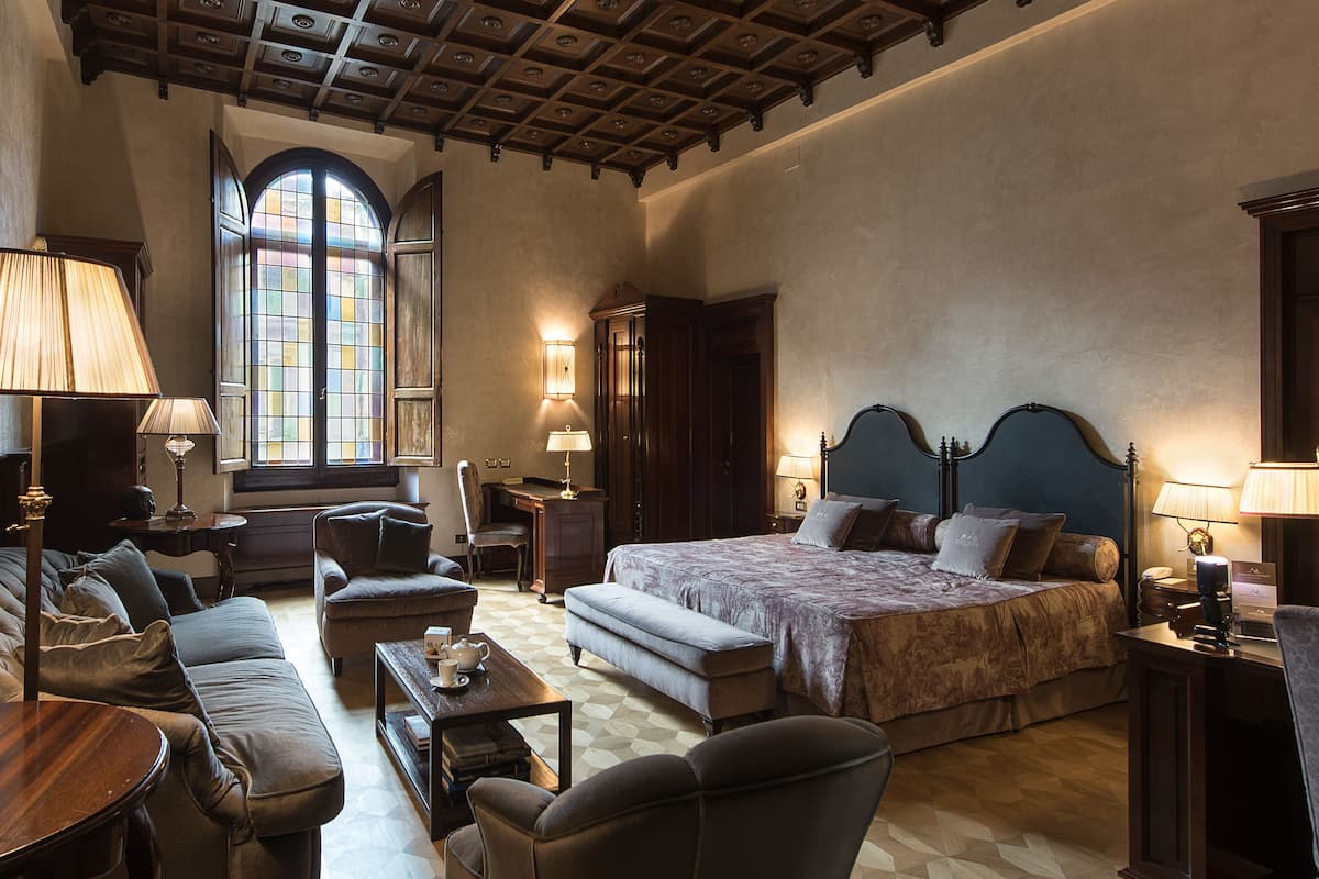 Ein geräumiges Schlafzimmer mit einem großen Bett, einer Holzbalkendecke, einem Fenster mit Buntglas und einer gemütlichen Sitzecke mit Sofa und Sesseln.