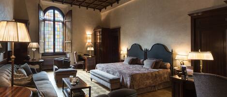 A spacious bedroom with a large bed, a wooden ceiling, a window with stained glass, and a cozy seating area with a sofa and armchairs.