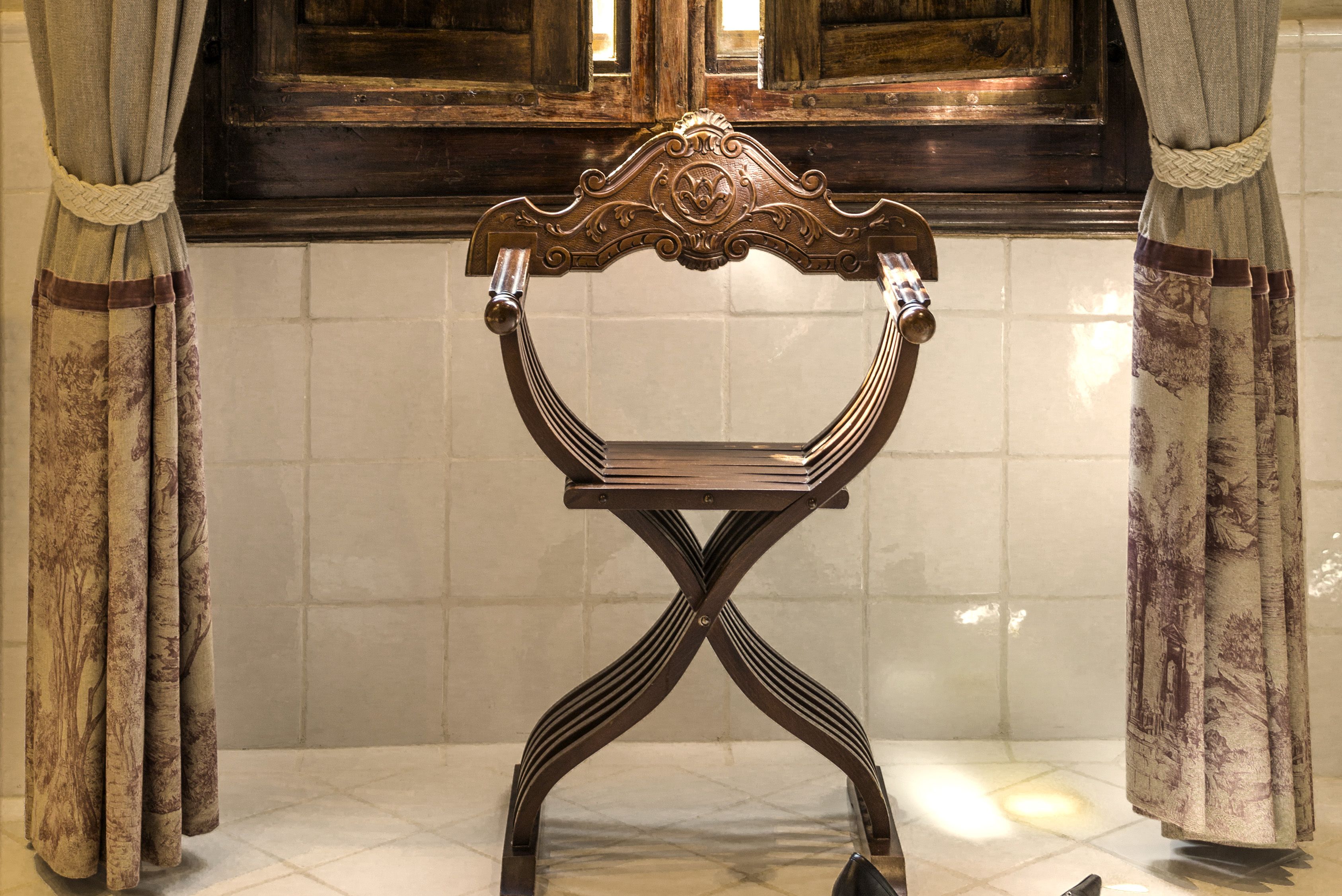 A wooden chair with an ornate backrest and a metal frame, positioned in a room with tiled walls and curtains.