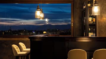 Eine Bar mit Blick auf die Stadt bei Dämmerung, mit zwei Barhocker und hängenden Pendelleuchten.