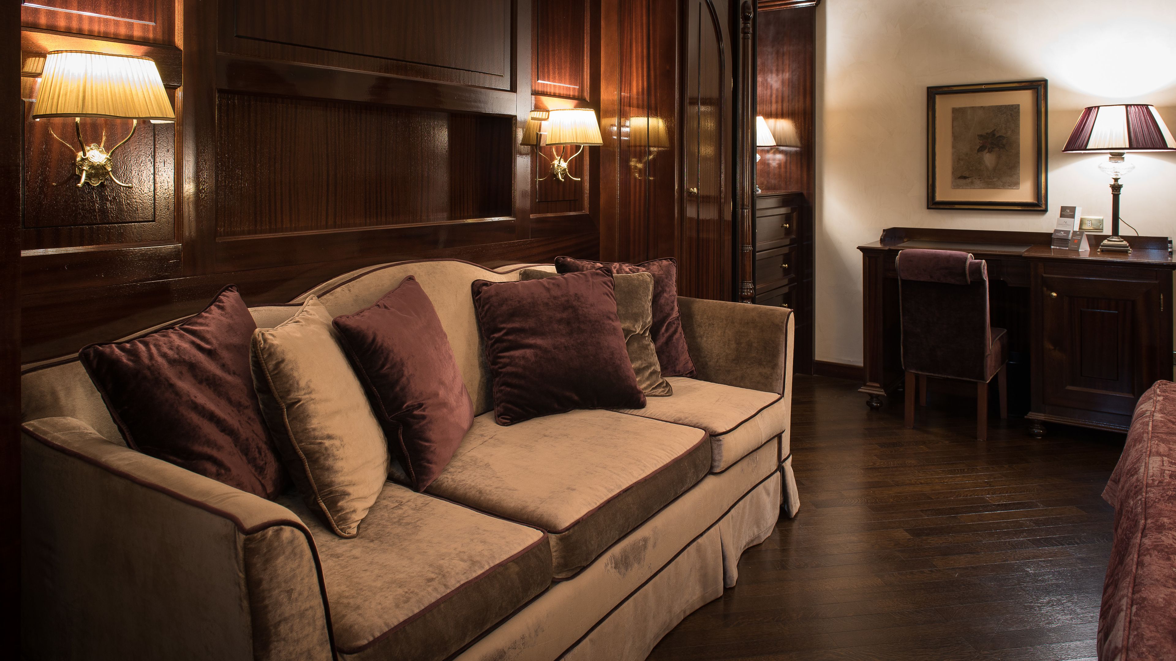 A cozy room with a wooden panel wall, a beige sofa with purple cushions, a desk with a chair, and wall-mounted lamps.