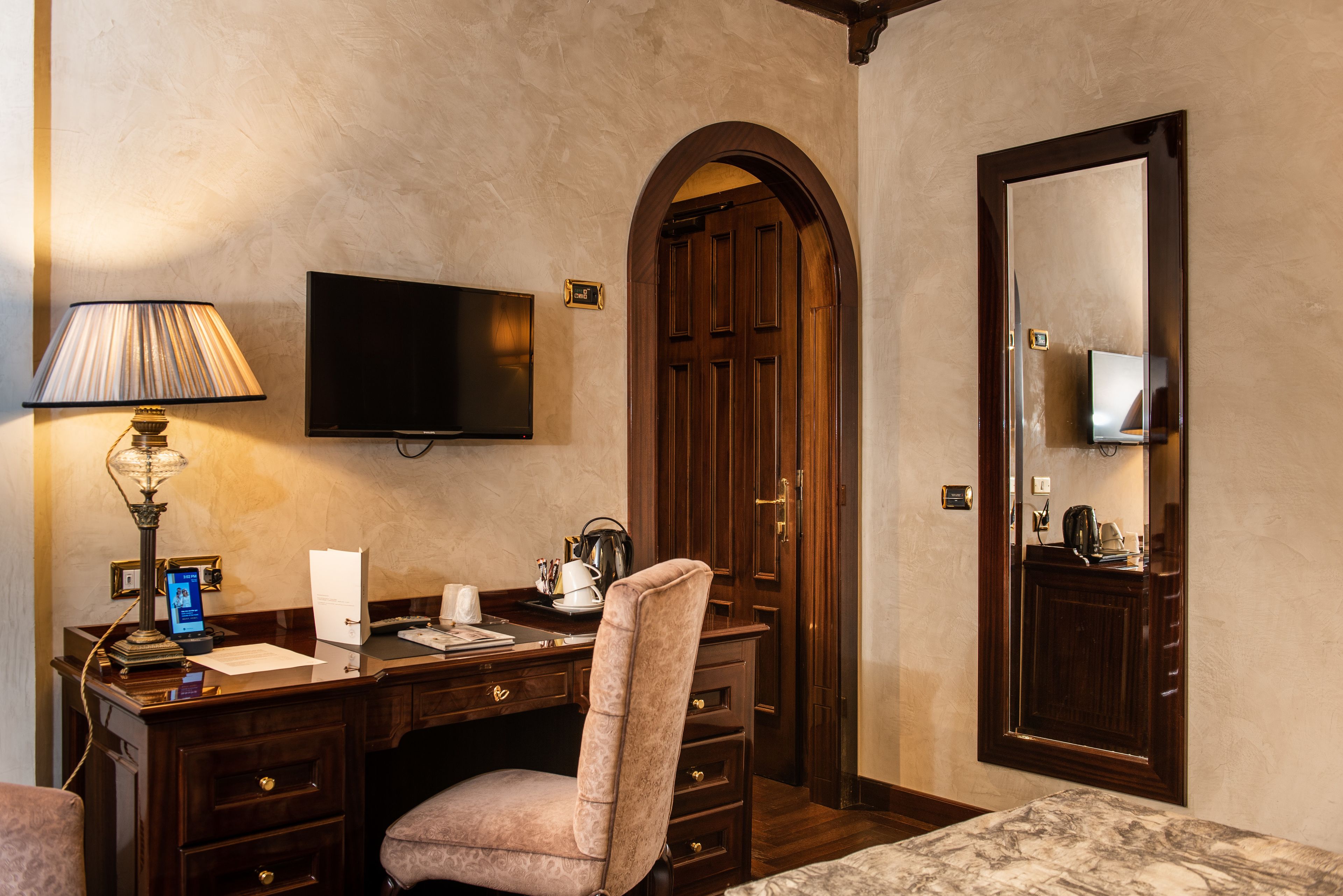 A hotel room with a wooden desk, a chair, a television mounted on the wall, and a large mirror.
