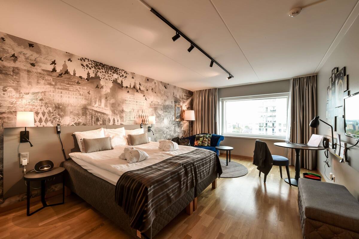 A hotel room with a large bed, a desk, a chair, and a view of the city through the window.