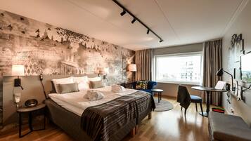 A hotel room with a large bed, a desk, a chair, and a view of the city through the window.