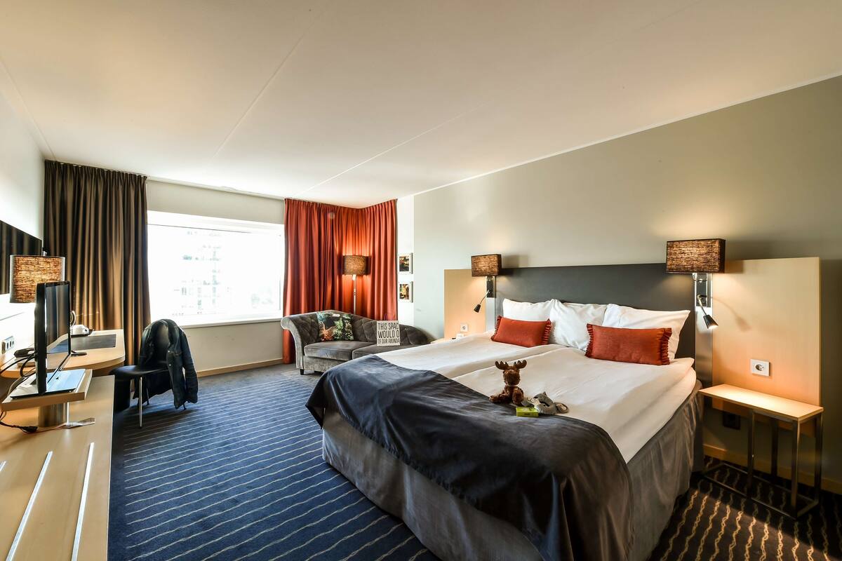 A hotel room with a large bed, a desk with a computer, a chair, a sofa, and a window with curtains.