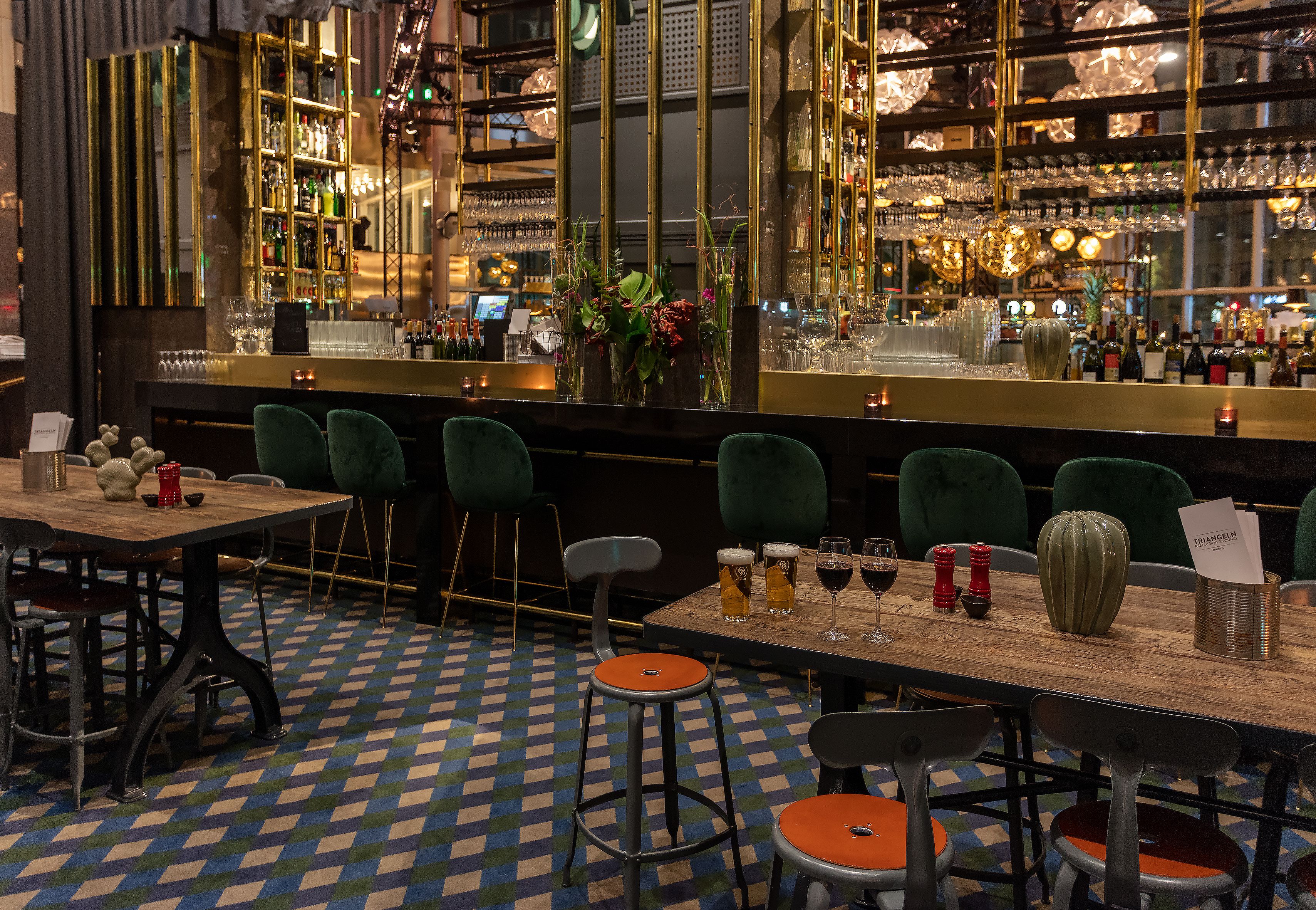 A bar area with checkered floor tiles, green upholstered chairs, and a wooden bar counter with glasses and bottles.