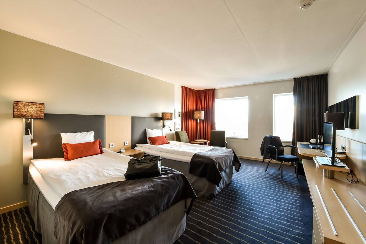 A hotel room with two beds, a desk, a chair, a TV, and a window with curtains.