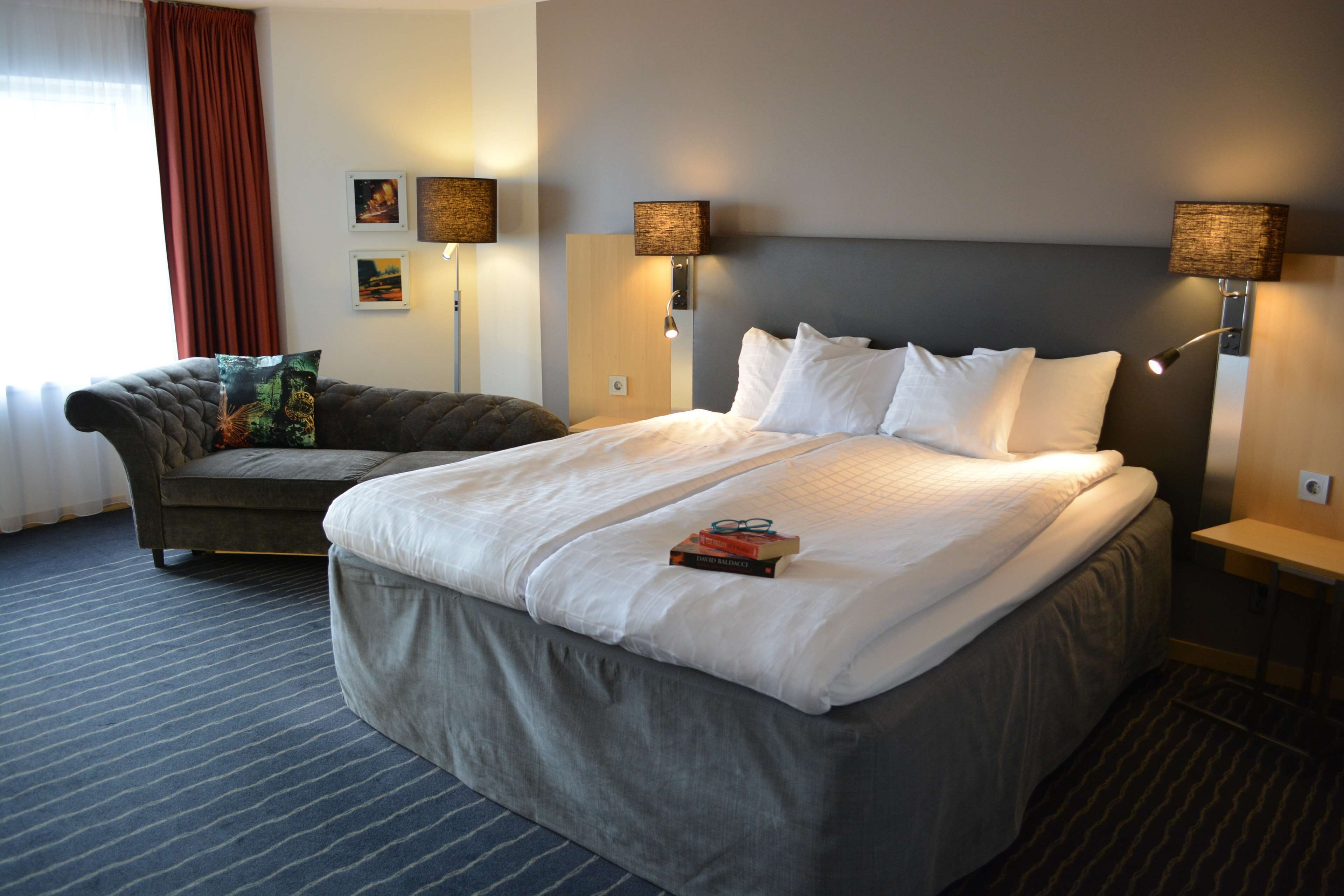 Ett hotellrum med en stor säng, en soffa, två vägglampor och ett fönster med gardiner.