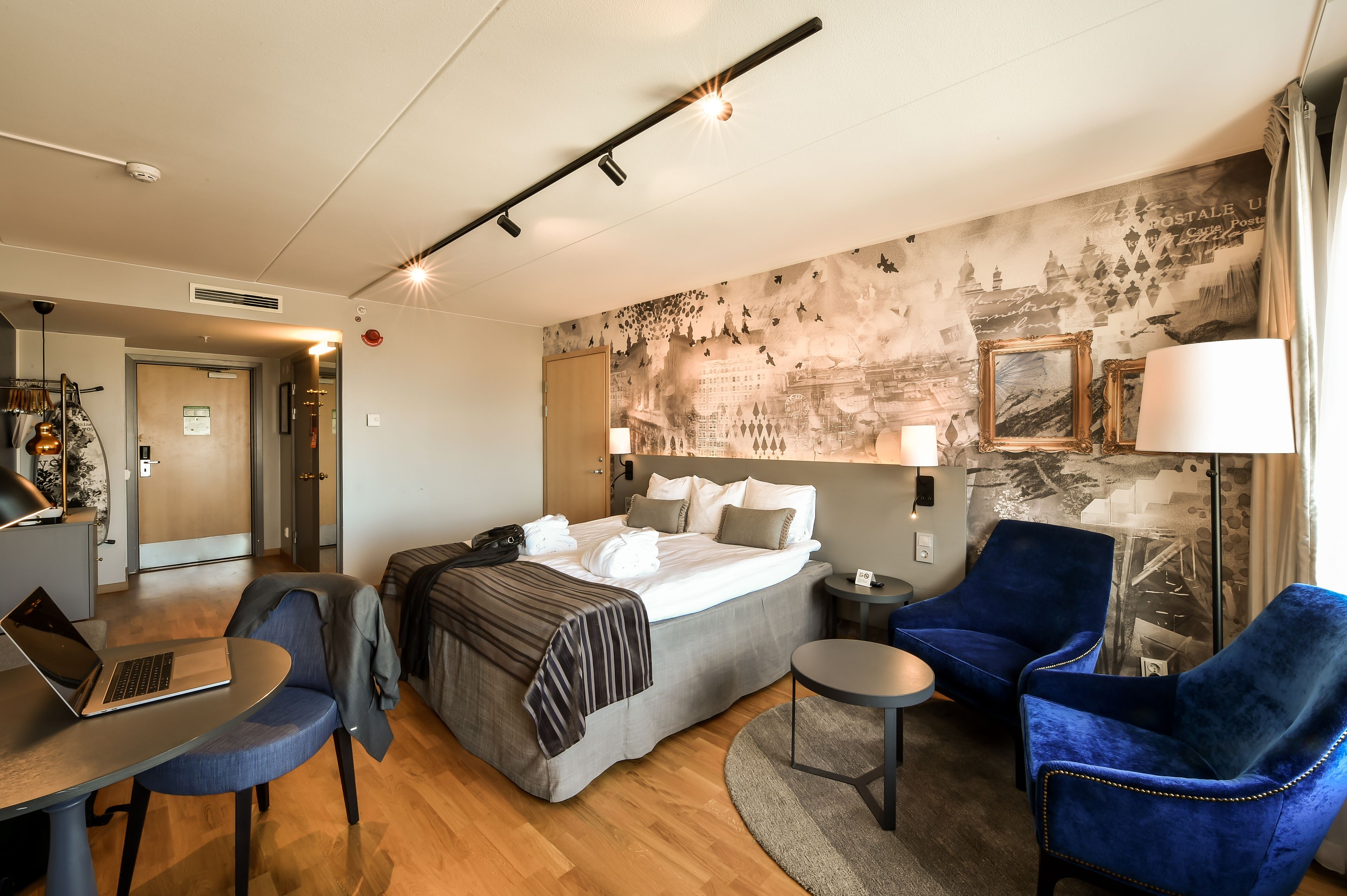 A hotel room with a bed, a desk with a laptop, two blue armchairs, a small round table, and a wall with a vintage cityscape mural.