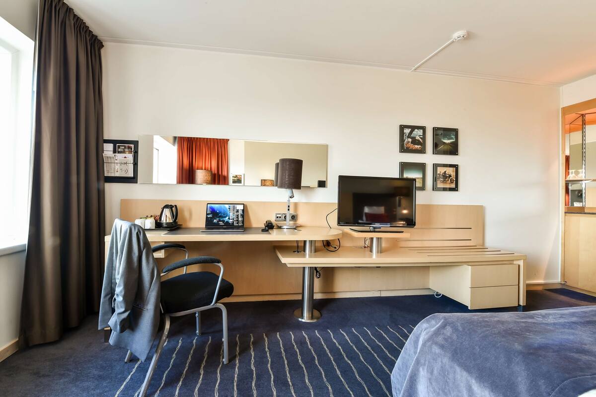 A hotel room with a bed, a desk with a computer, a chair, a TV, a lamp, and a window with curtains.
