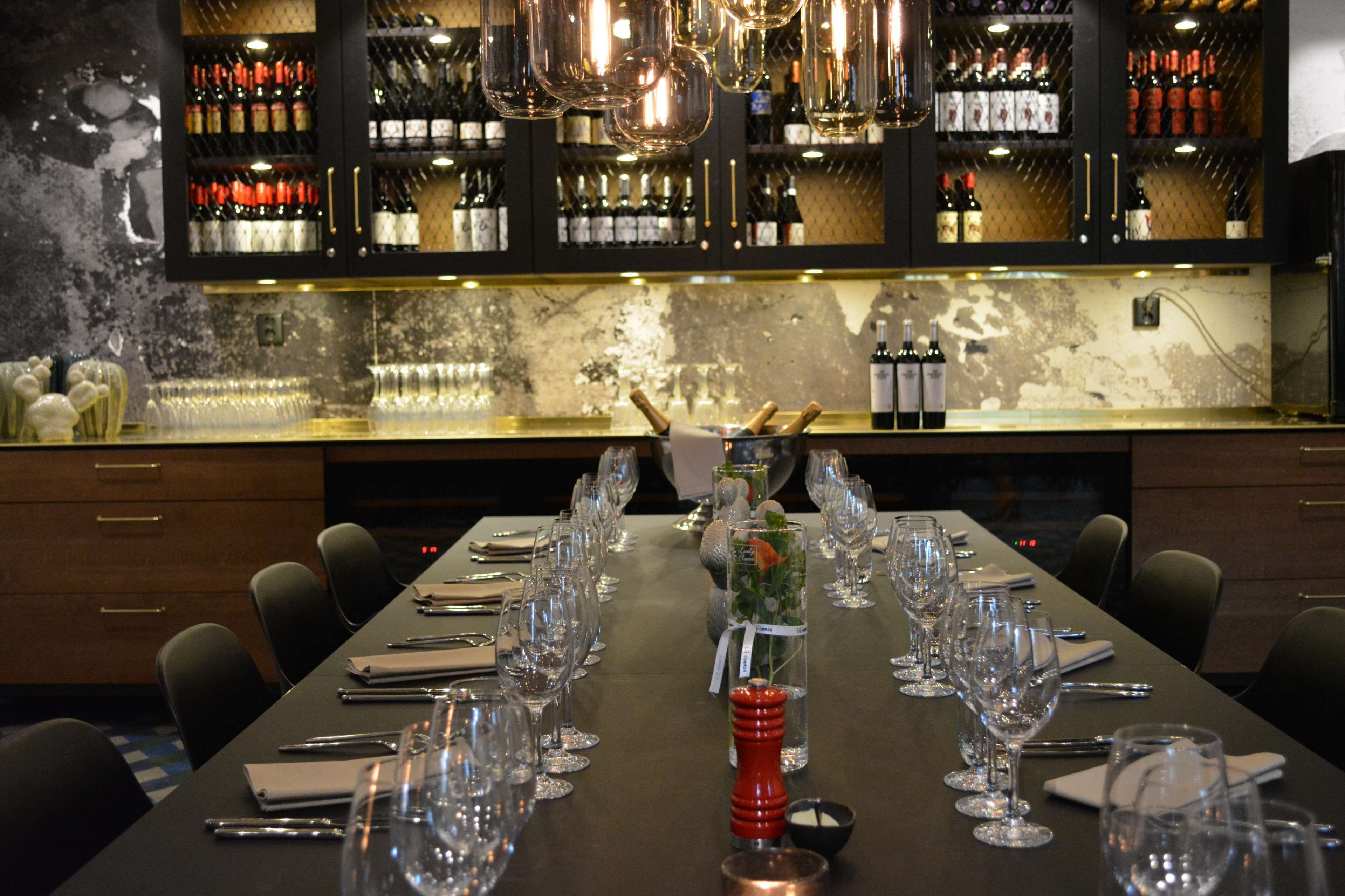 Una larga mesa de comedor dispuesta con copas de vino, salero y pimentero, y un centro de mesa decorativo. Al fondo de la mesa, se ve una botellera con varias botellas.