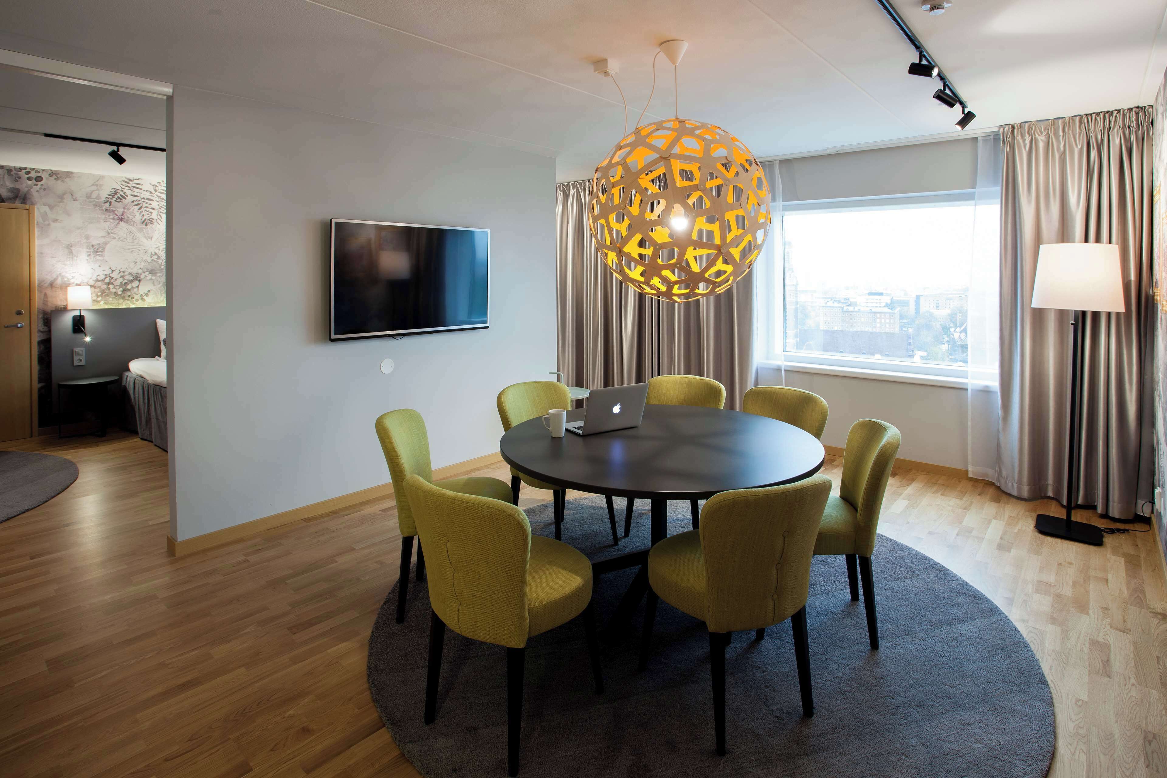 A modern meeting room with a round table, six chairs, a laptop, a TV, and a large decorative light fixture.