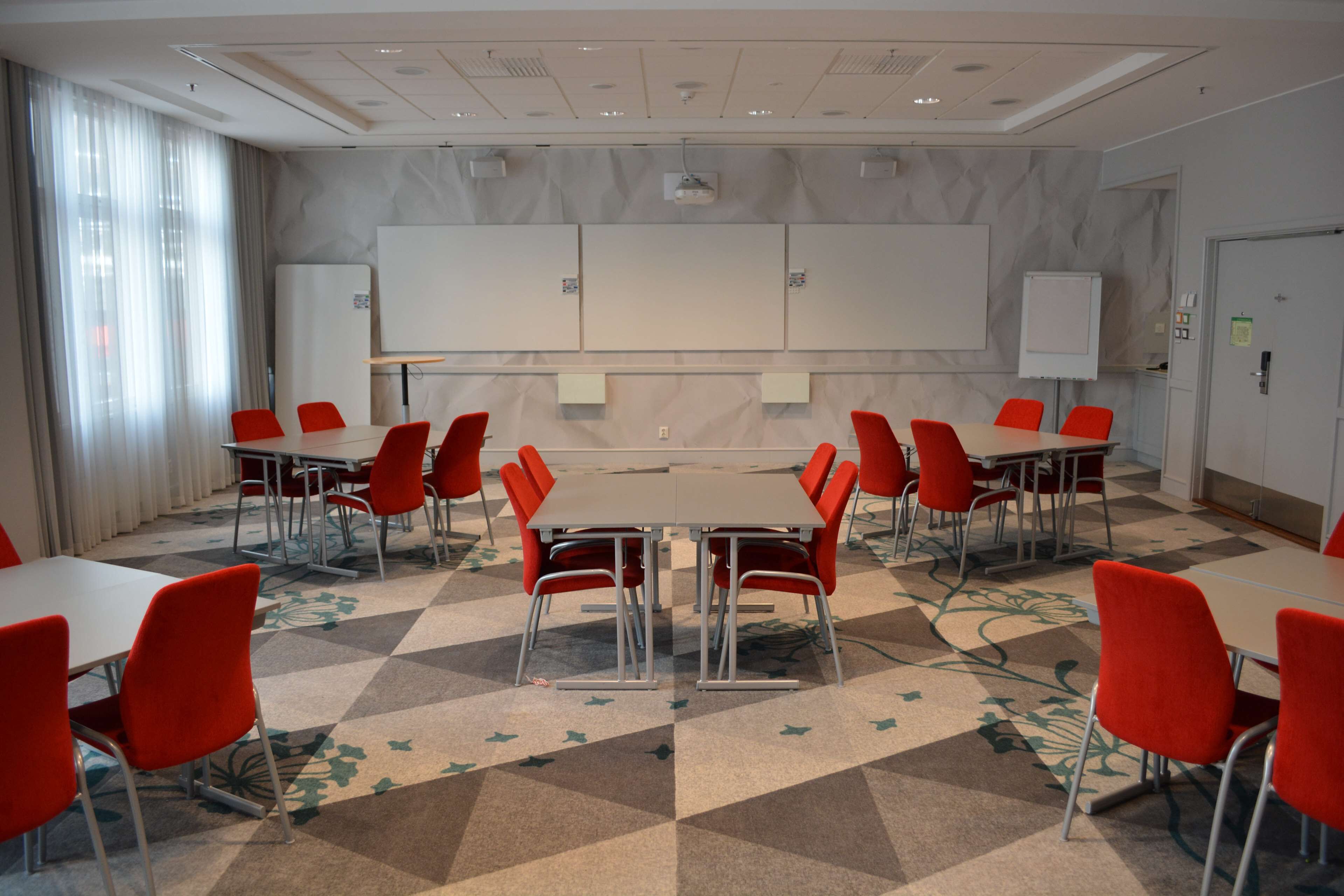 Une salle de conférence dotée d’une grande table entourée de chaises rouges, de tableaux blancs et d’un tapis à motifs.