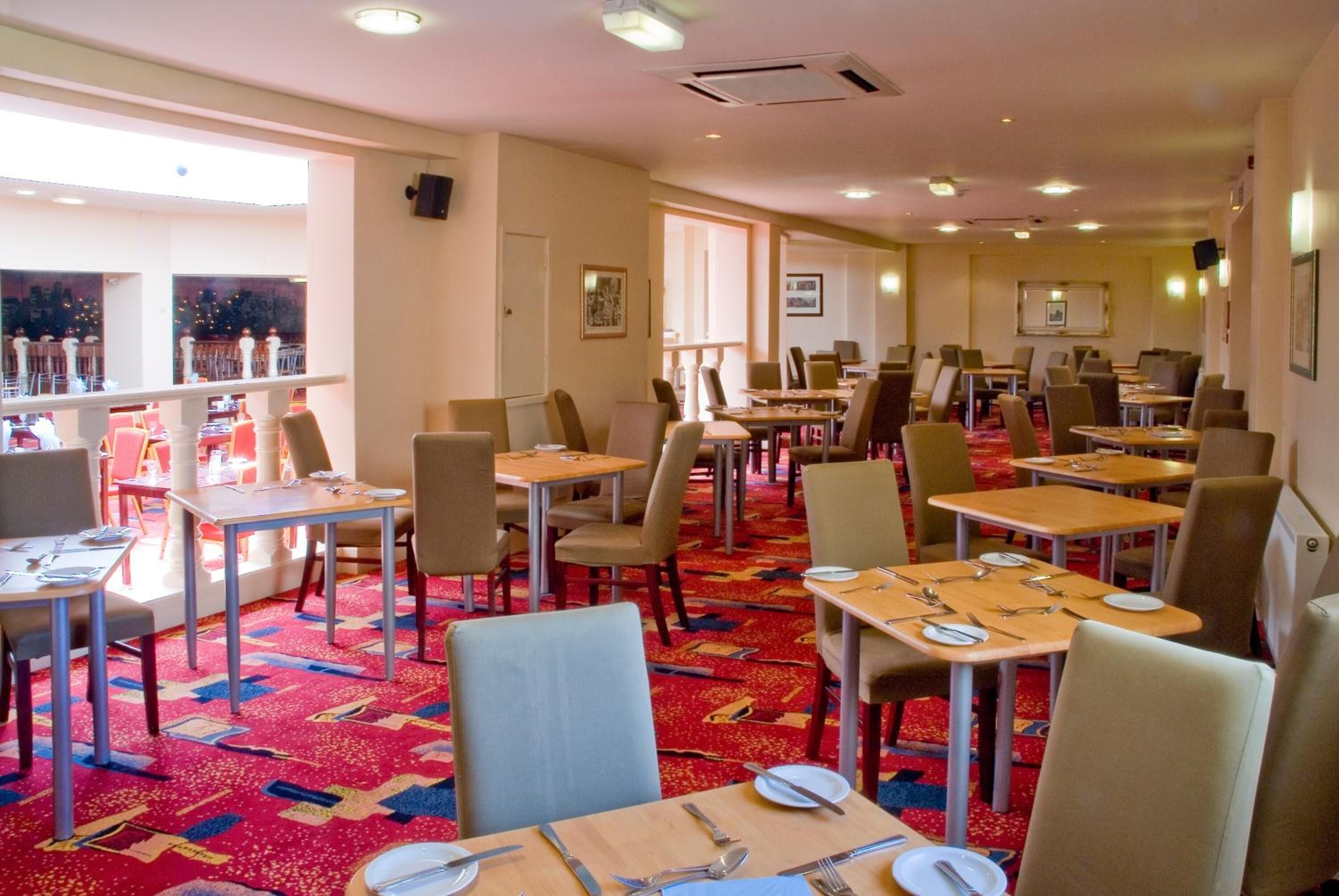 Ein Hotelrestaurant mit mehreren Tischen und Stühlen, einem gemusterten Teppich und Blick durch große Fenster.