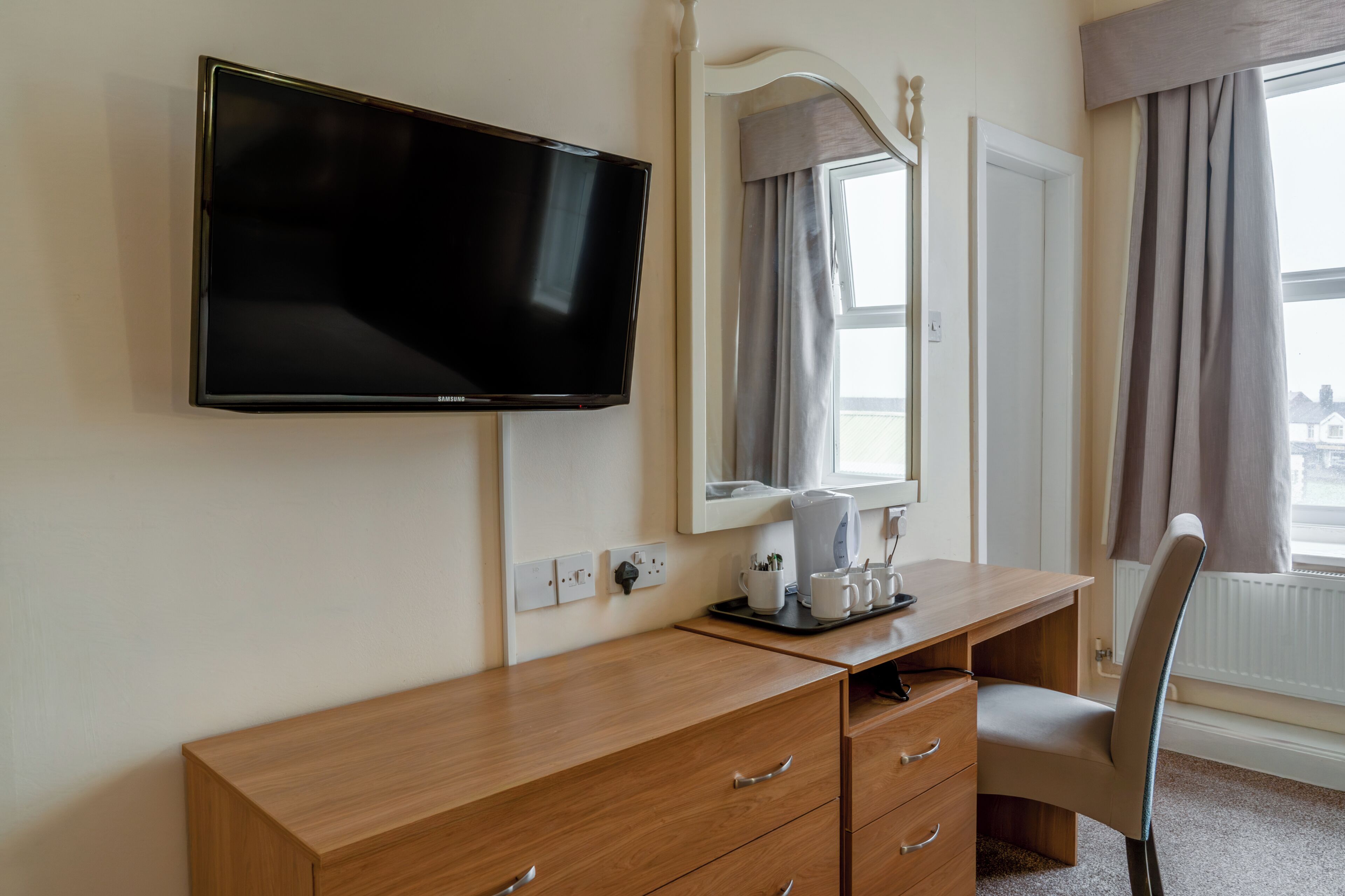 A hotel room with a flat-screen TV, a wooden desk, a chair, a mirror, a window with curtains, and a radiator.