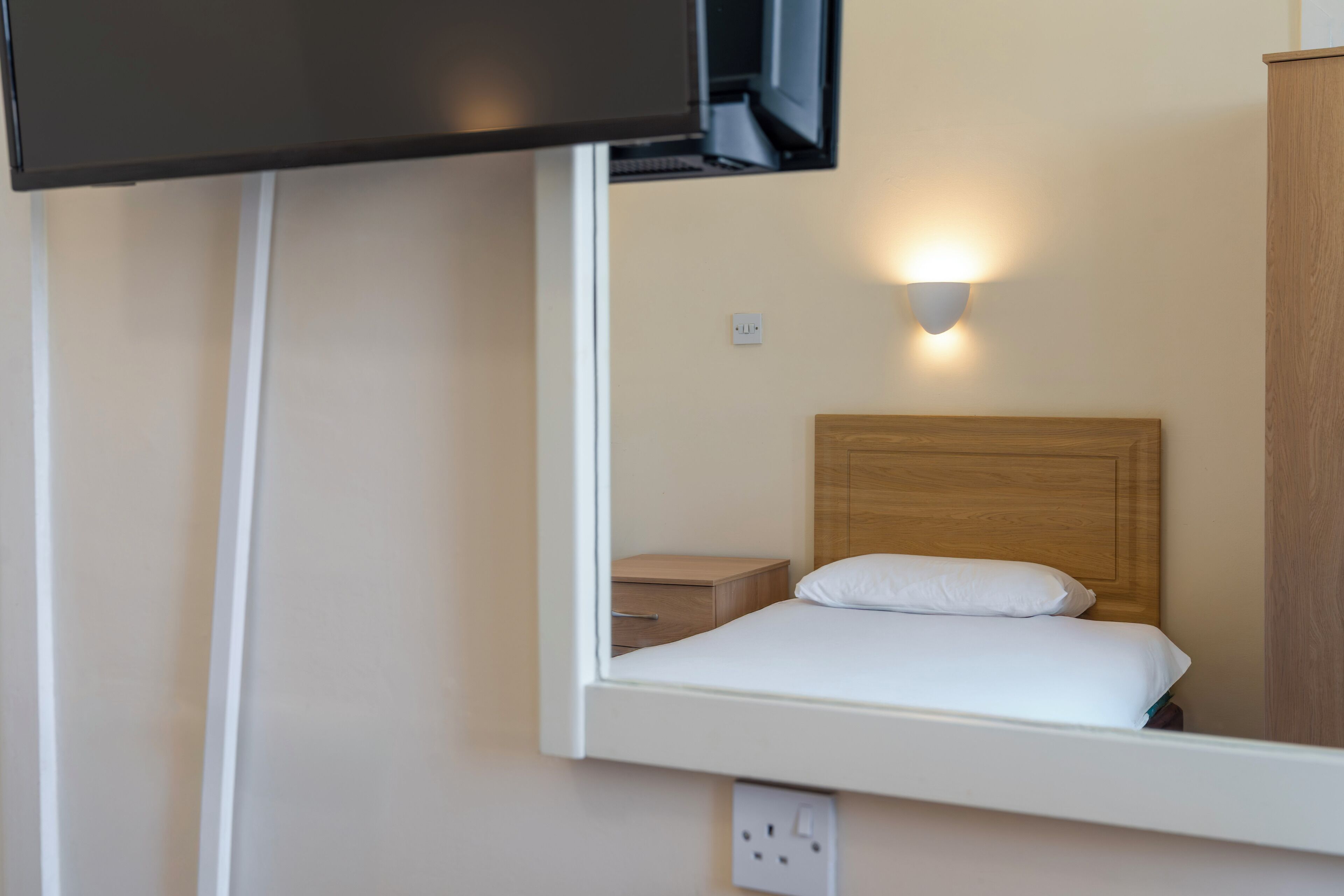 Een hotelkamer met een bed, een nachtkastje, een tv boven het bed, een wandlamp en een houten garderobe.