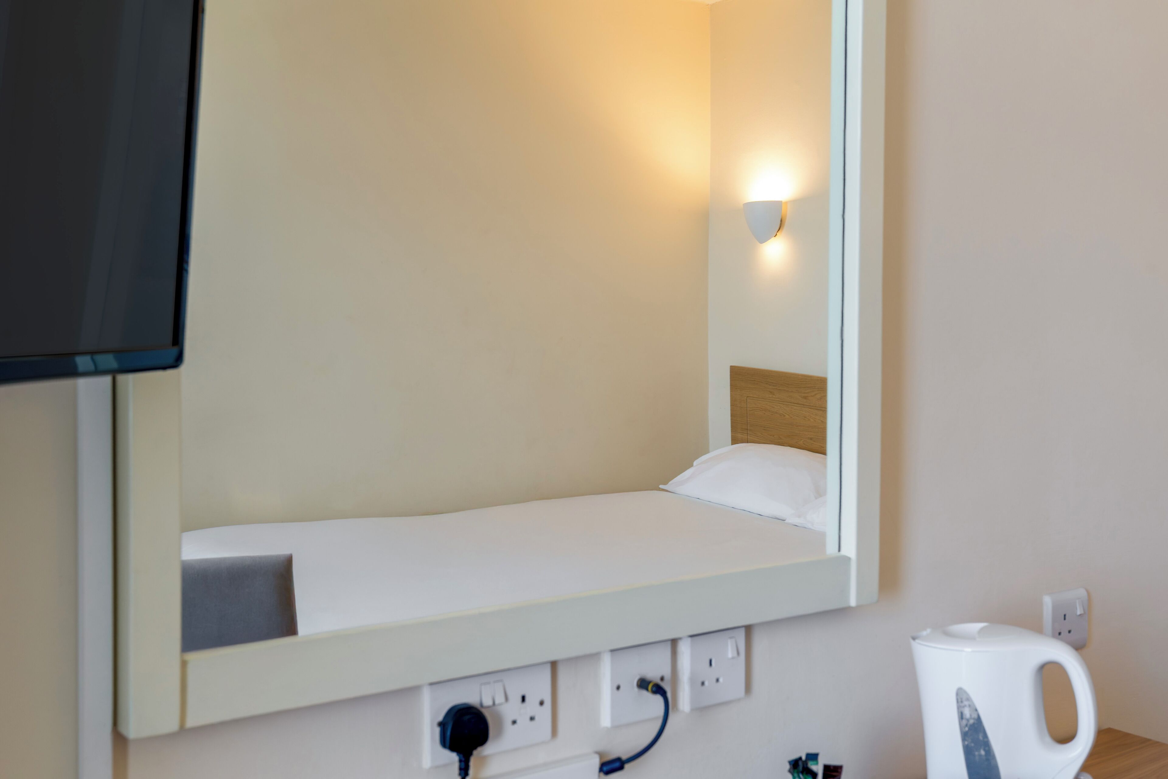A hotel room with a bed, a TV mounted above it, a bedside table with a kettle, and a wall socket.