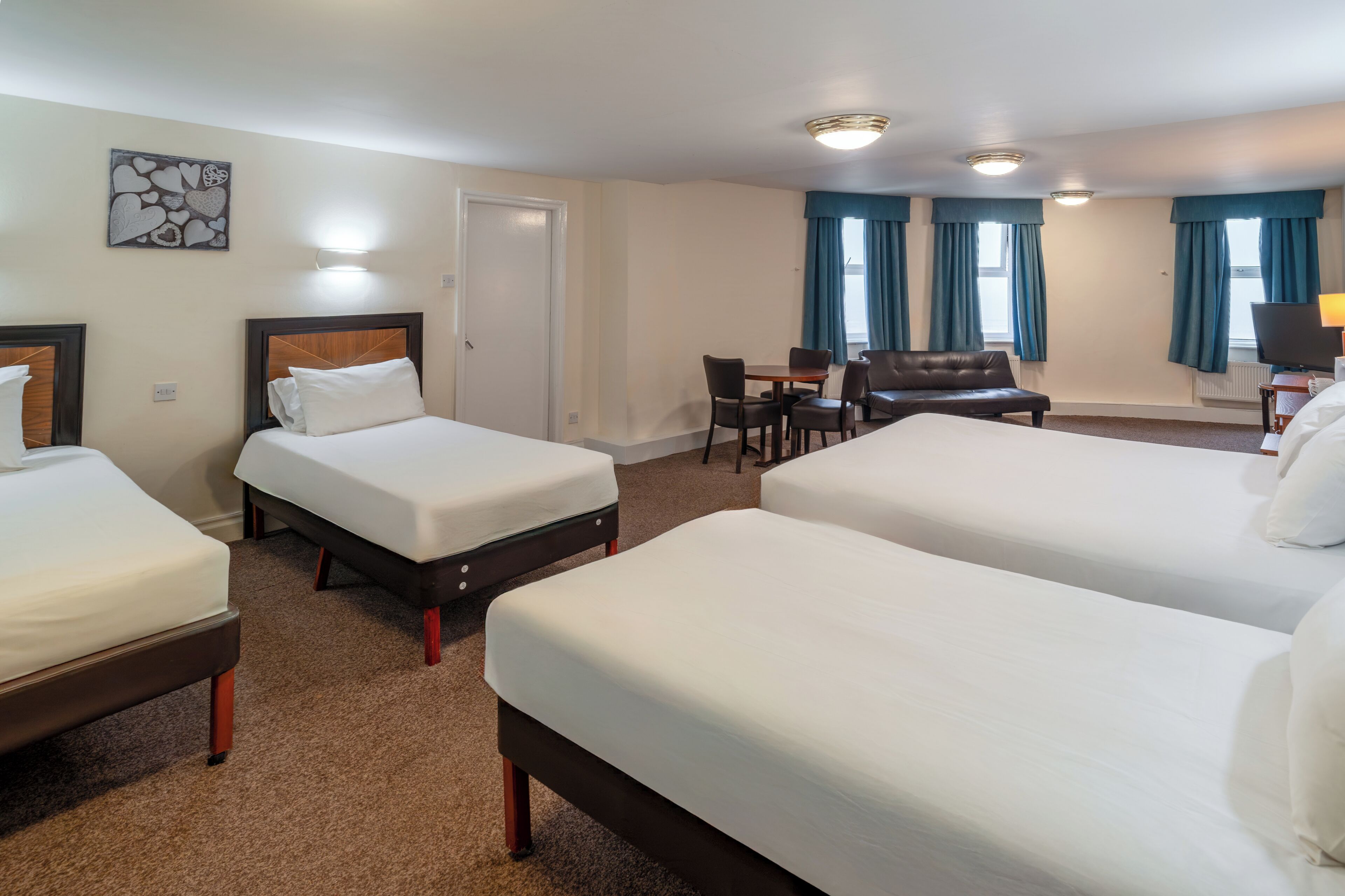 A hotel room with three beds, a dining table with chairs, a TV, and a desk.