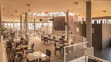 A spacious restaurant with high ceilings, multiple tables and chairs, and a view of the city through large windows.