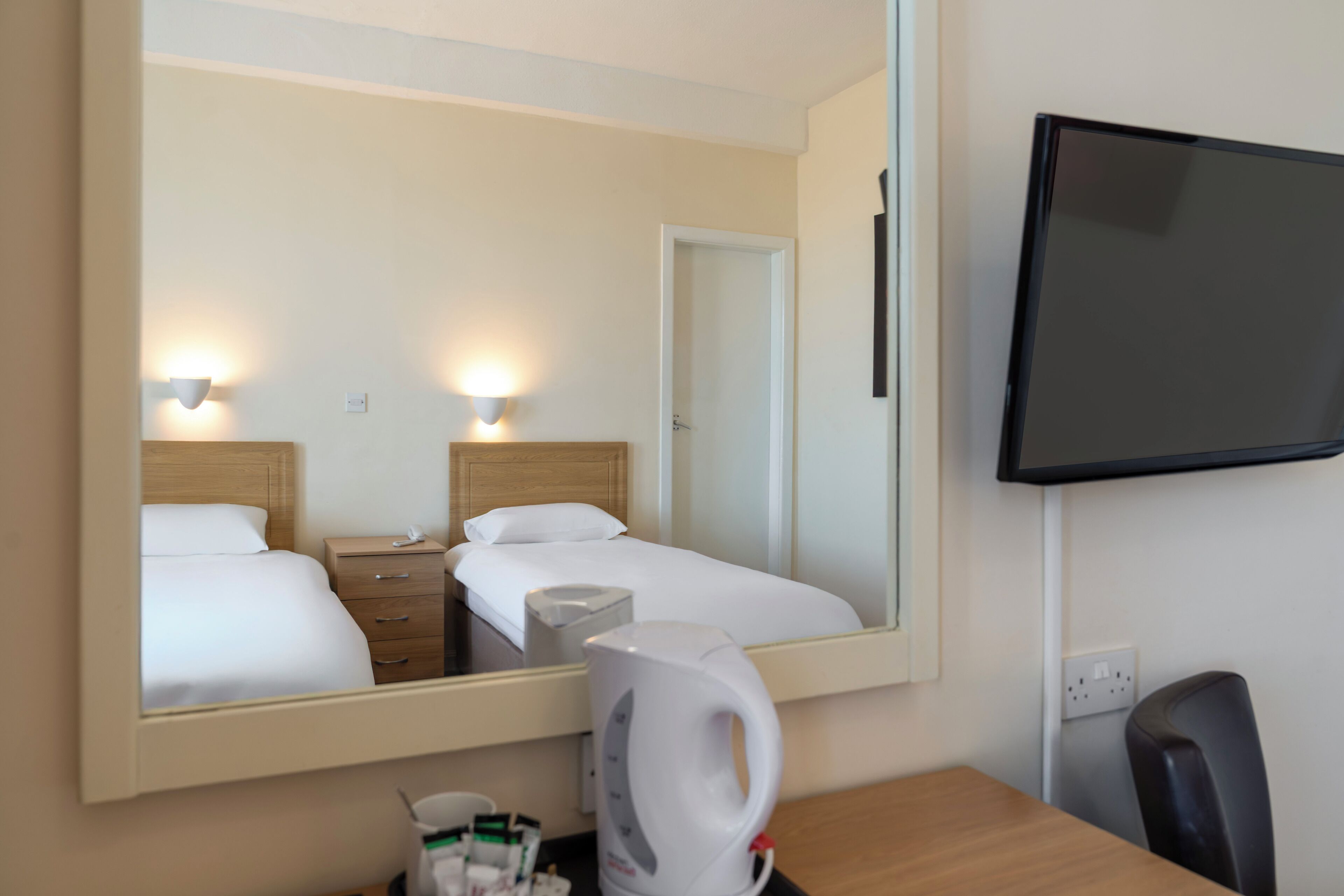 Una habitación de hotel con cama, mesita de noche, espejo, televisor y cafetera.