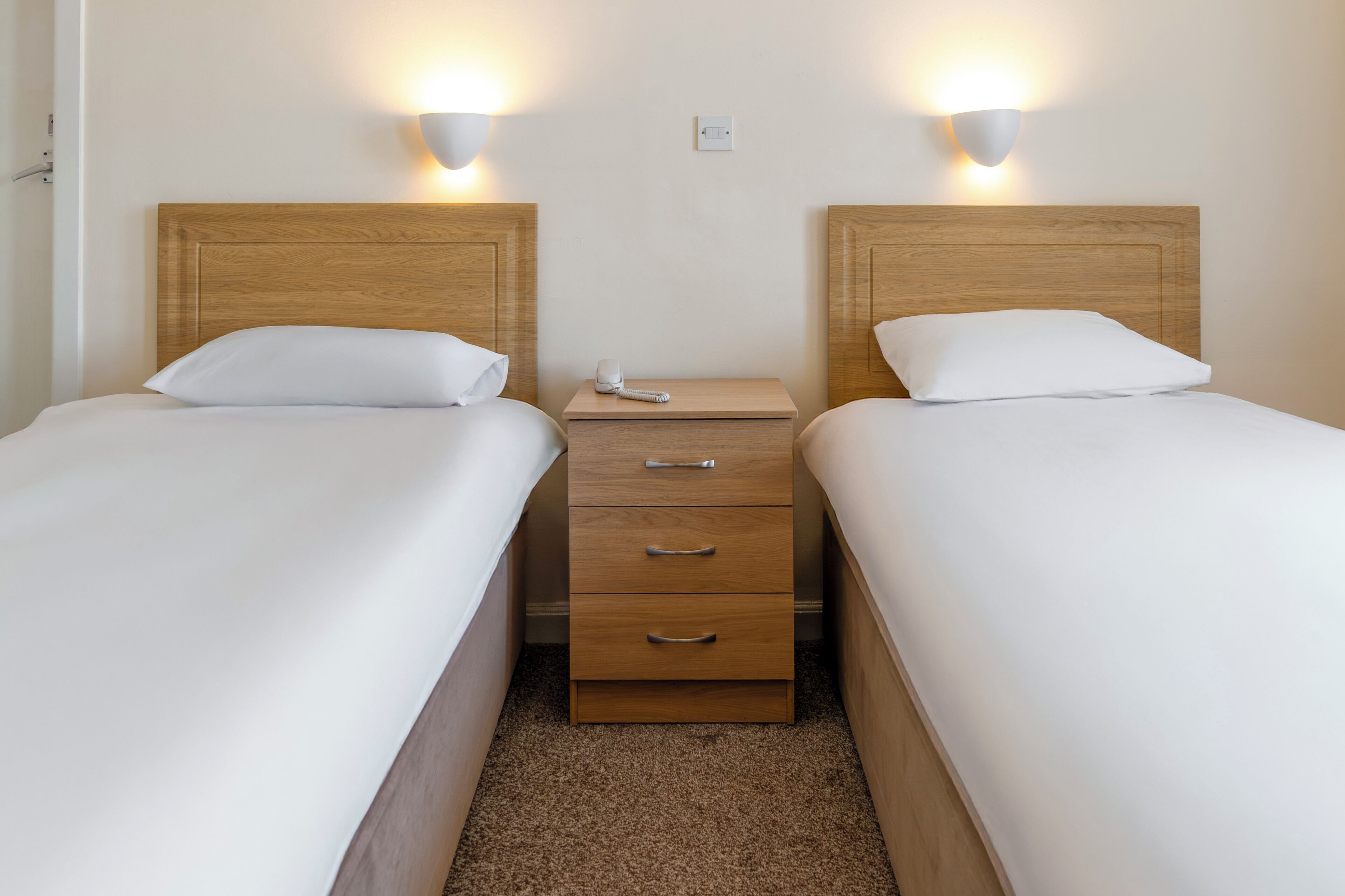 Quarto de hotel com duas camas de solteiro, uma mesa de cabeceira de madeira com gavetas e luminárias fixadas na parede.