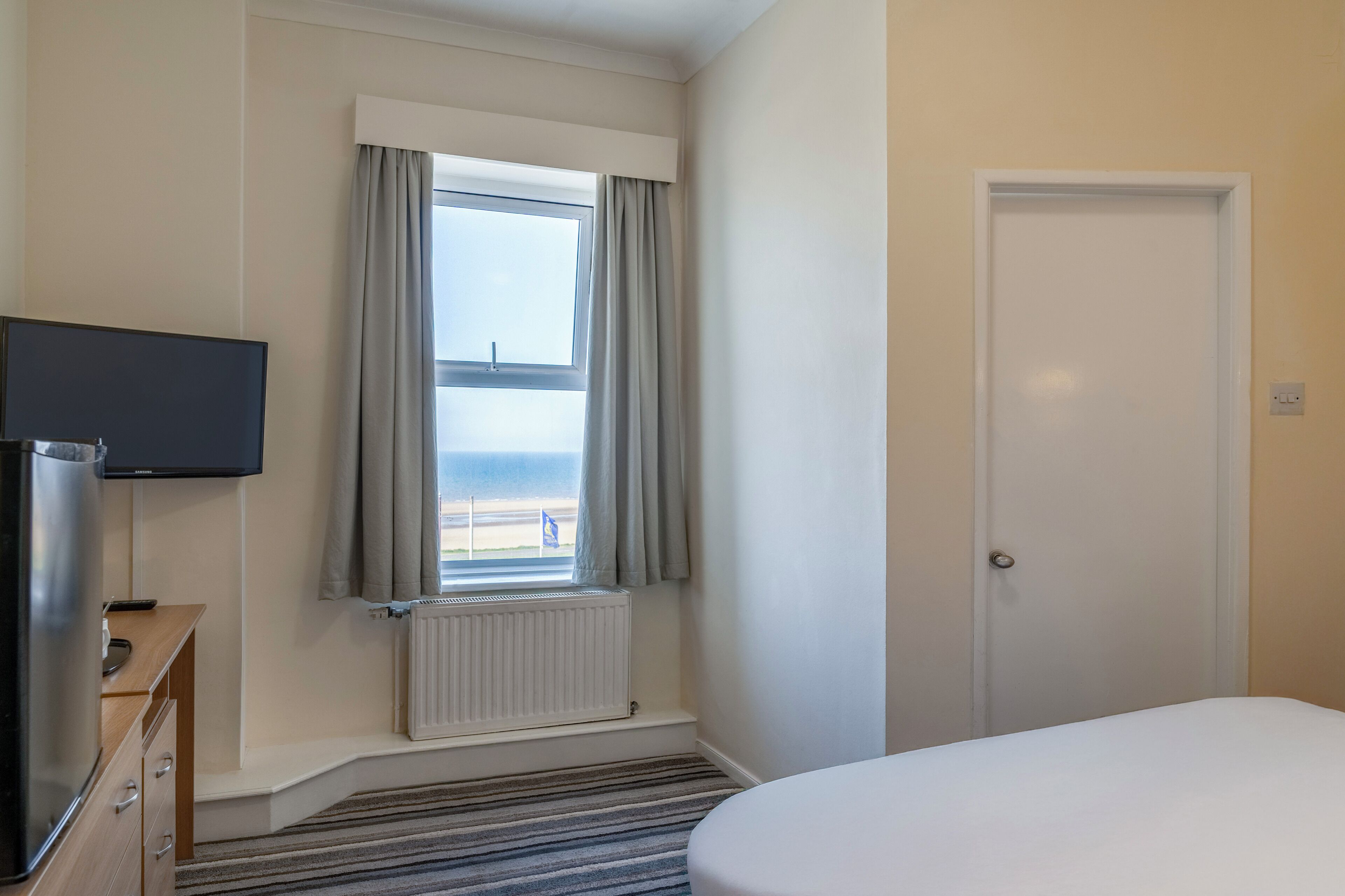 A hotel room with a bed, a television, a window with curtains, a radiator, and a door.