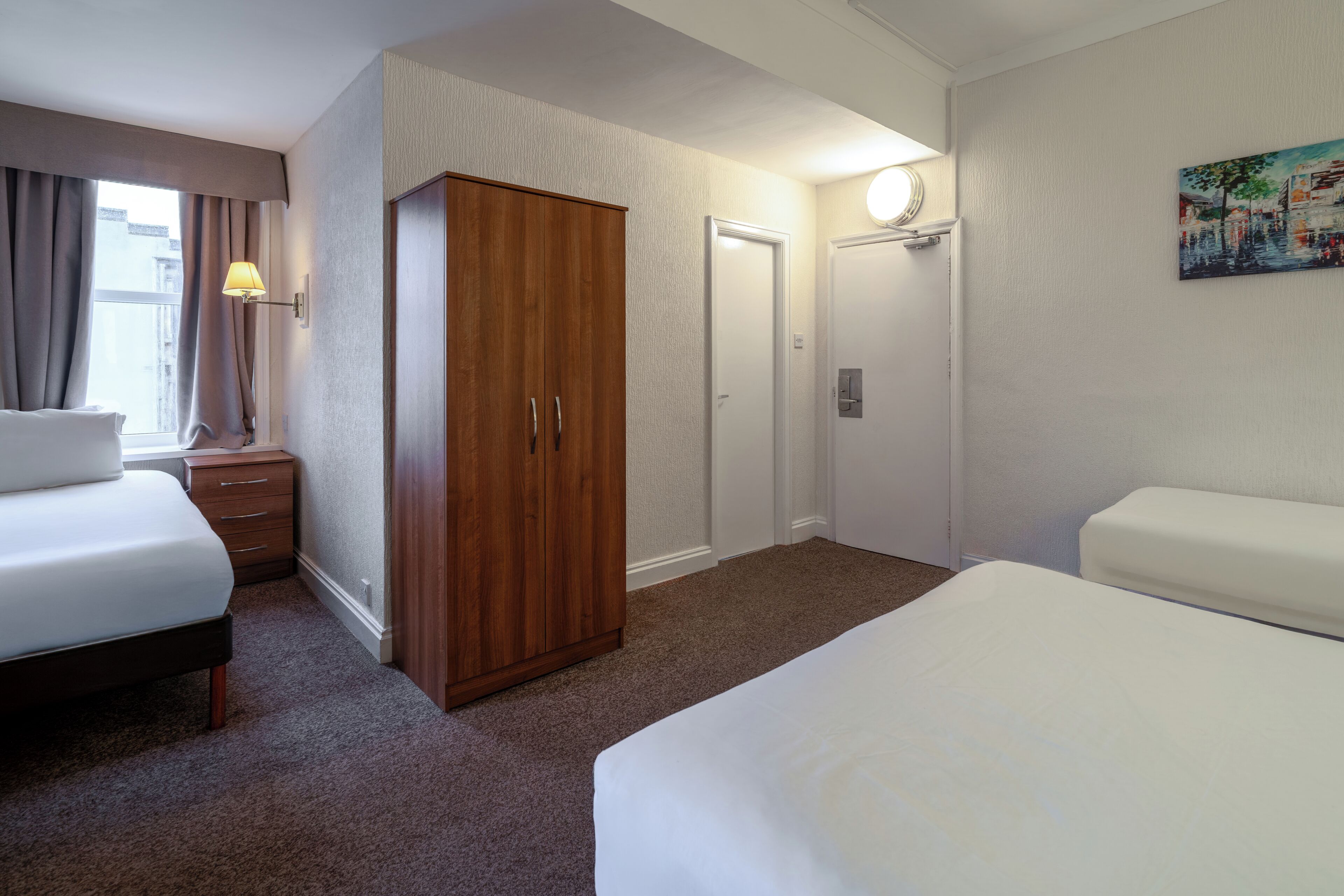 A hotel room with a bed, a wardrobe, a nightstand, a lamp, a door, and a painting on the wall.