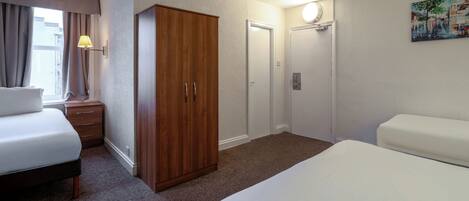 A hotel room with a bed, a wardrobe, a nightstand, a lamp, a door, and a painting on the wall.