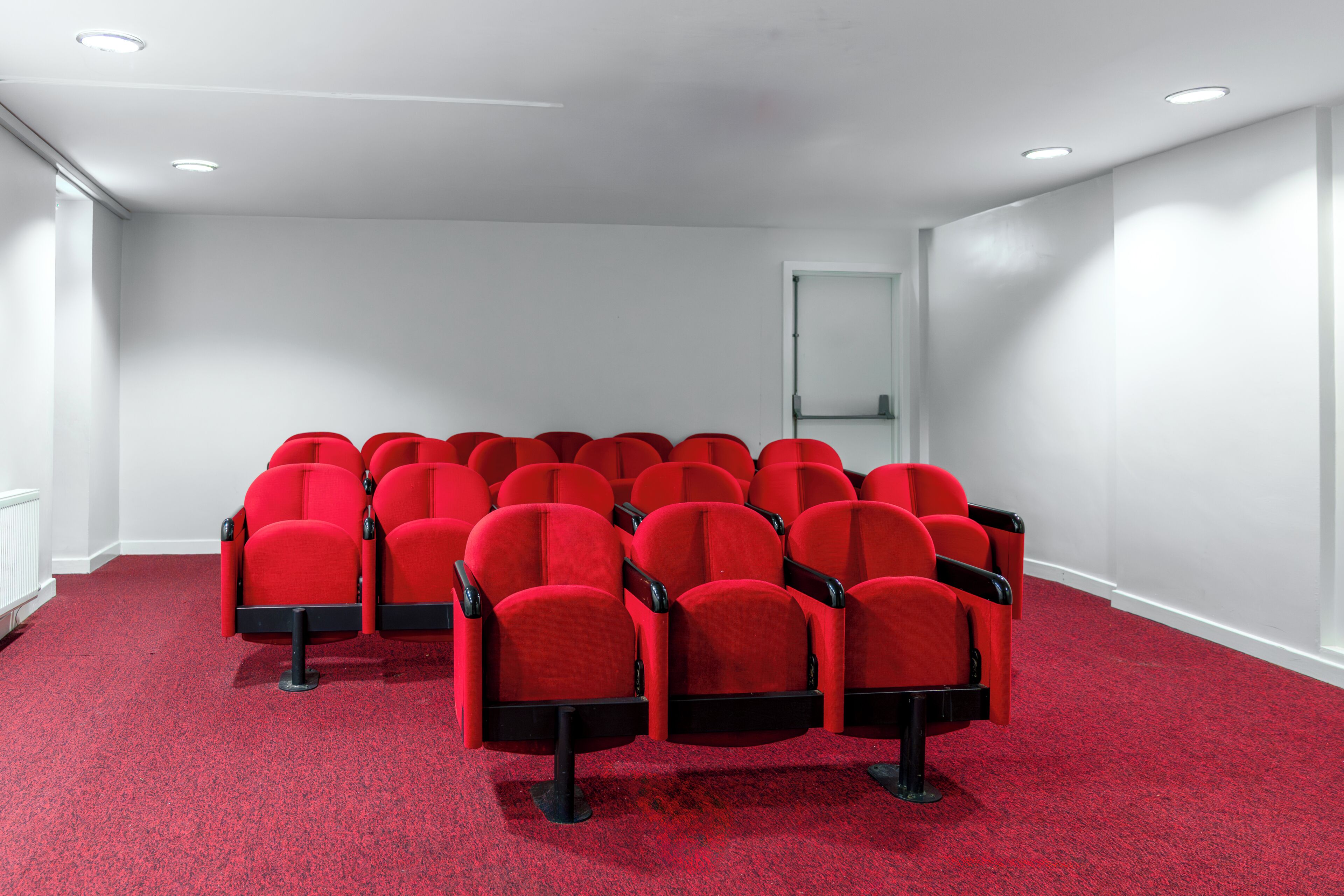 A conference room with rows of red chairs, white walls, and a carpeted floor.