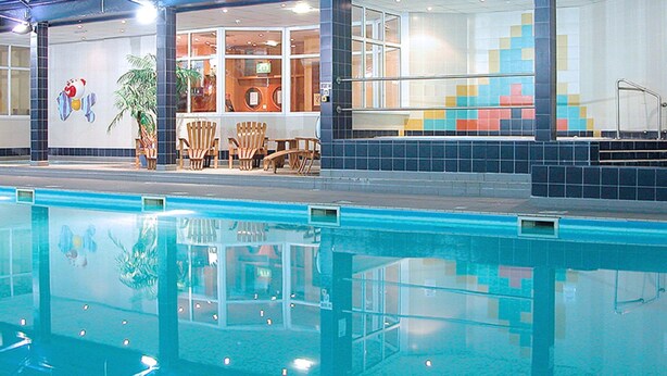 An indoor swimming pool with clear water, wooden chairs, and a tiled wall with a colorful pattern. - Norbreck Castle Hotel Blackpool (Blackpool)