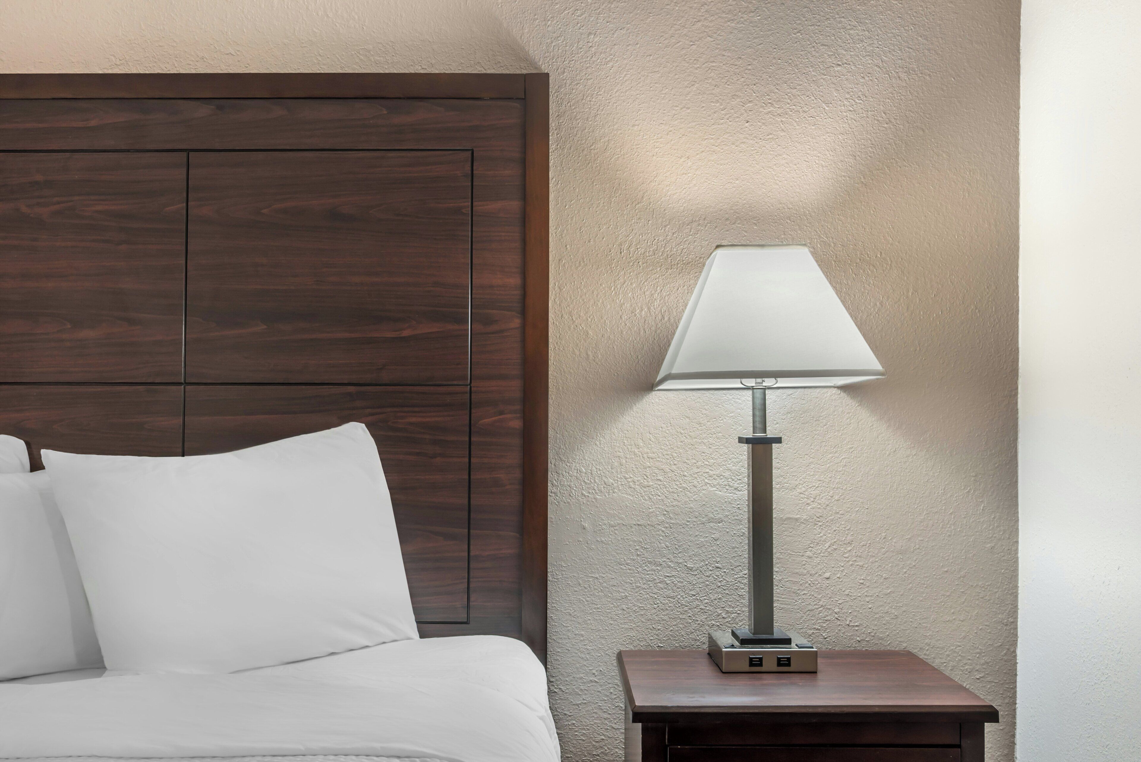 A bed with white bedding, a wooden headboard, a bedside table with a lamp, and a textured wall.